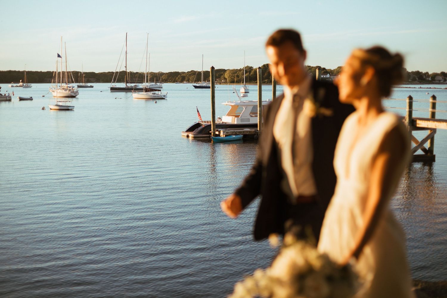 Bride and groom walking hand in hand with sailboats and serene harbor visible in background.