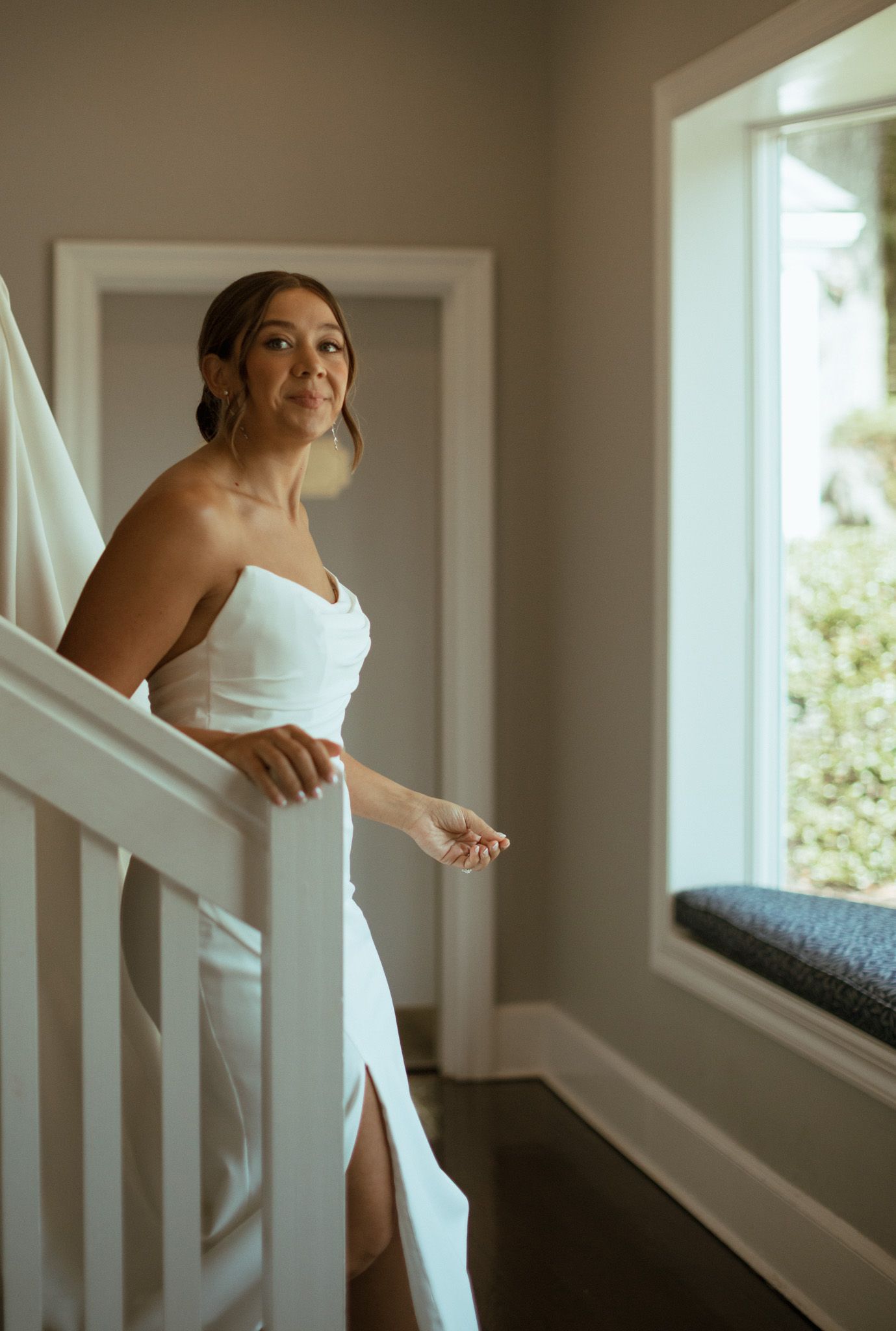 Bride in strapless white wedding gown standing on staircase near window before ceremony.