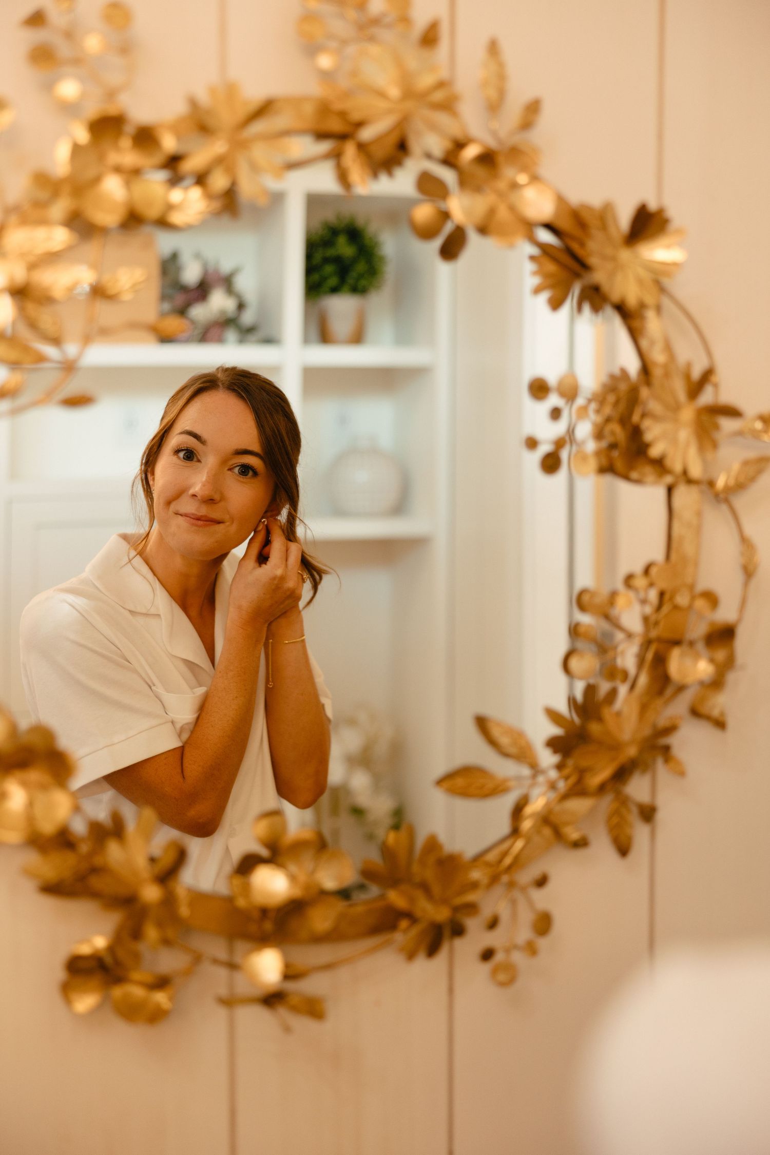 Bride in white robe putting on earrings reflected in ornate gold floral framed mirror.