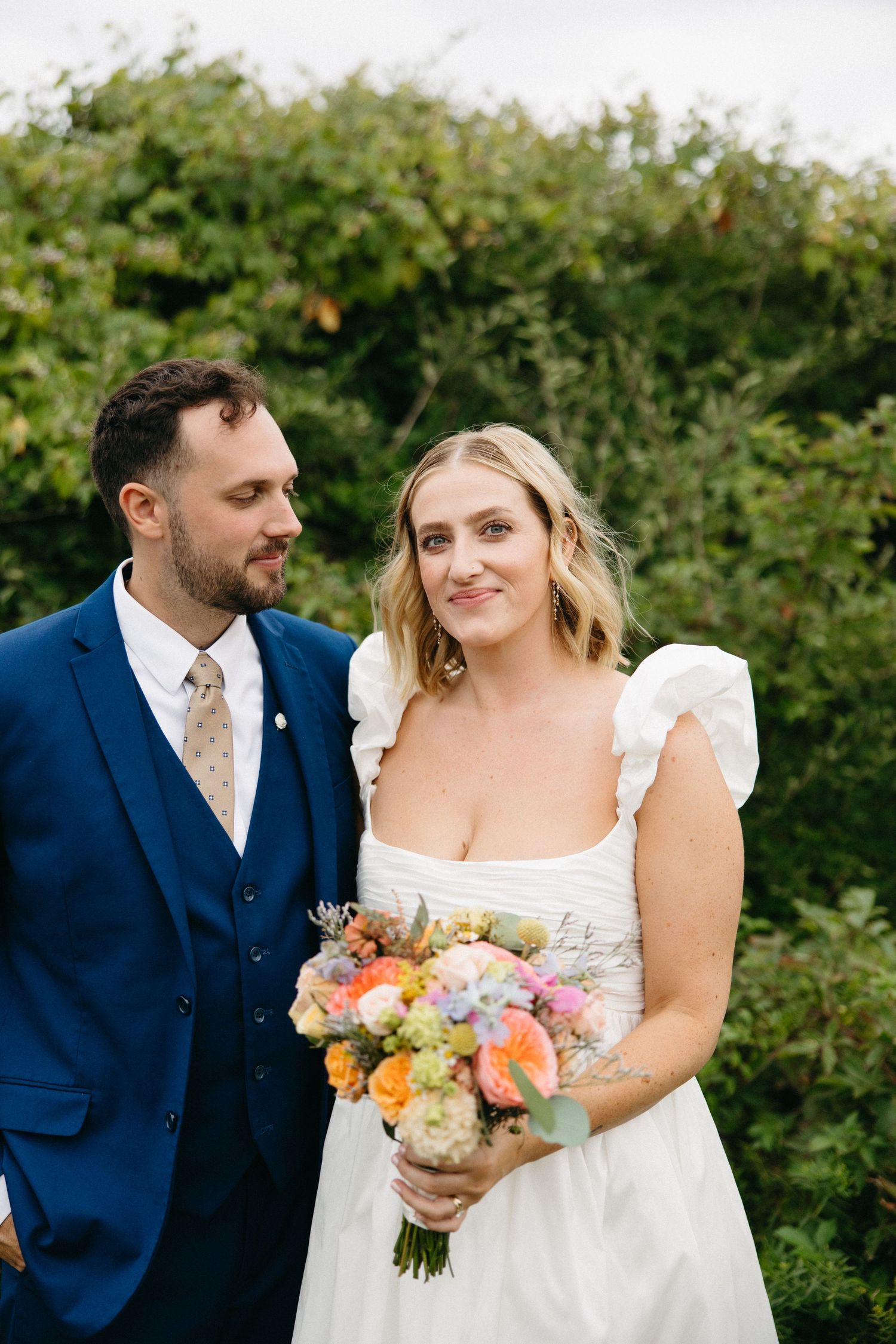 Bride and groom pose together outdoors, she holds a colorful bouquet, he wears a navy suit with floral tie.