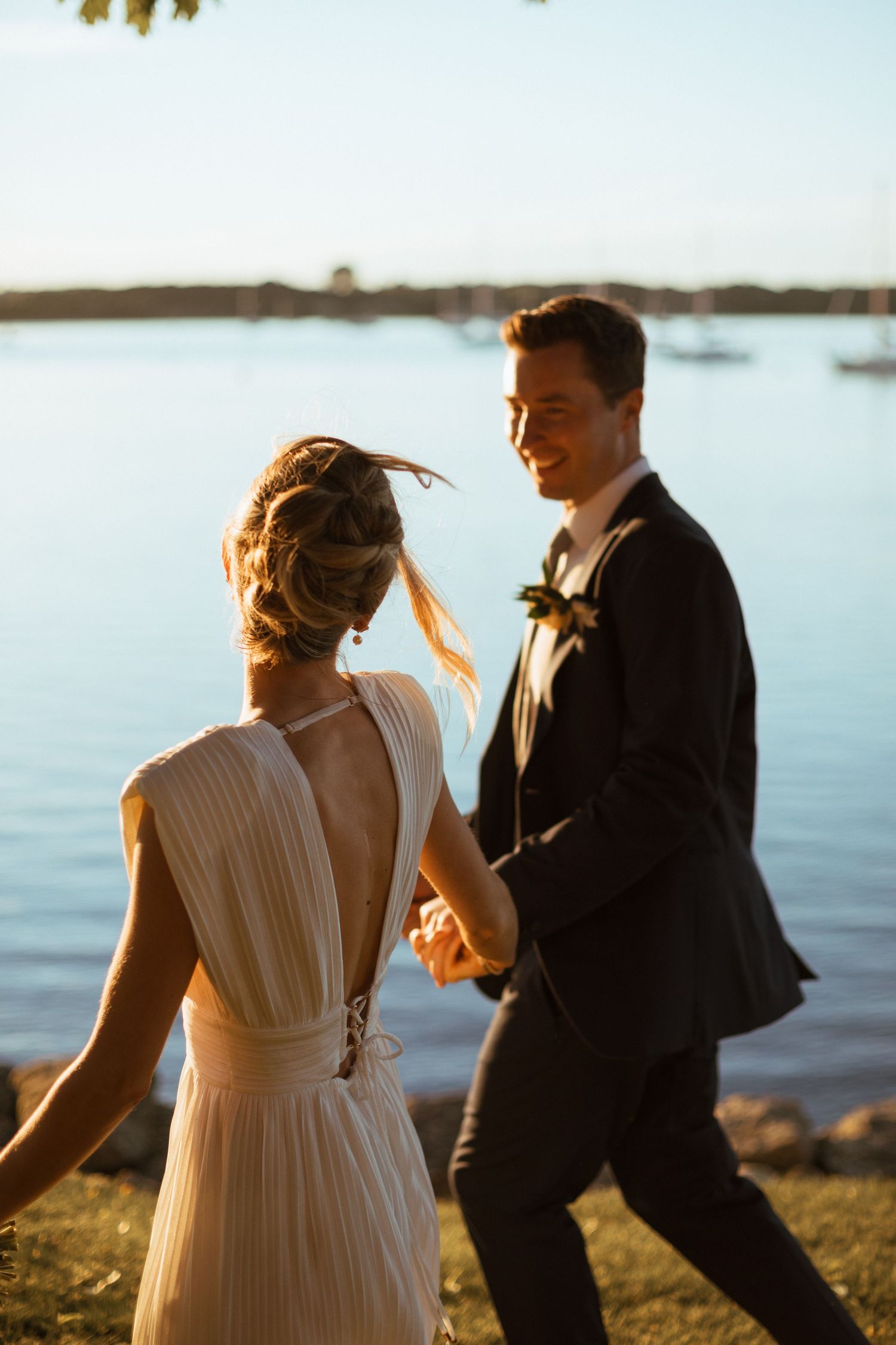 Smiling groom leading bride by the hand along waterfront during beautiful golden hour wedding photos.