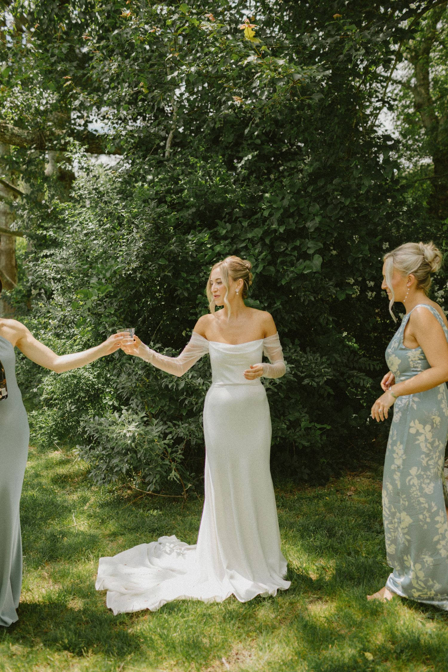 Bride in off-shoulder gown dancing joyfully with bridesmaids in sage dresses outdoors.