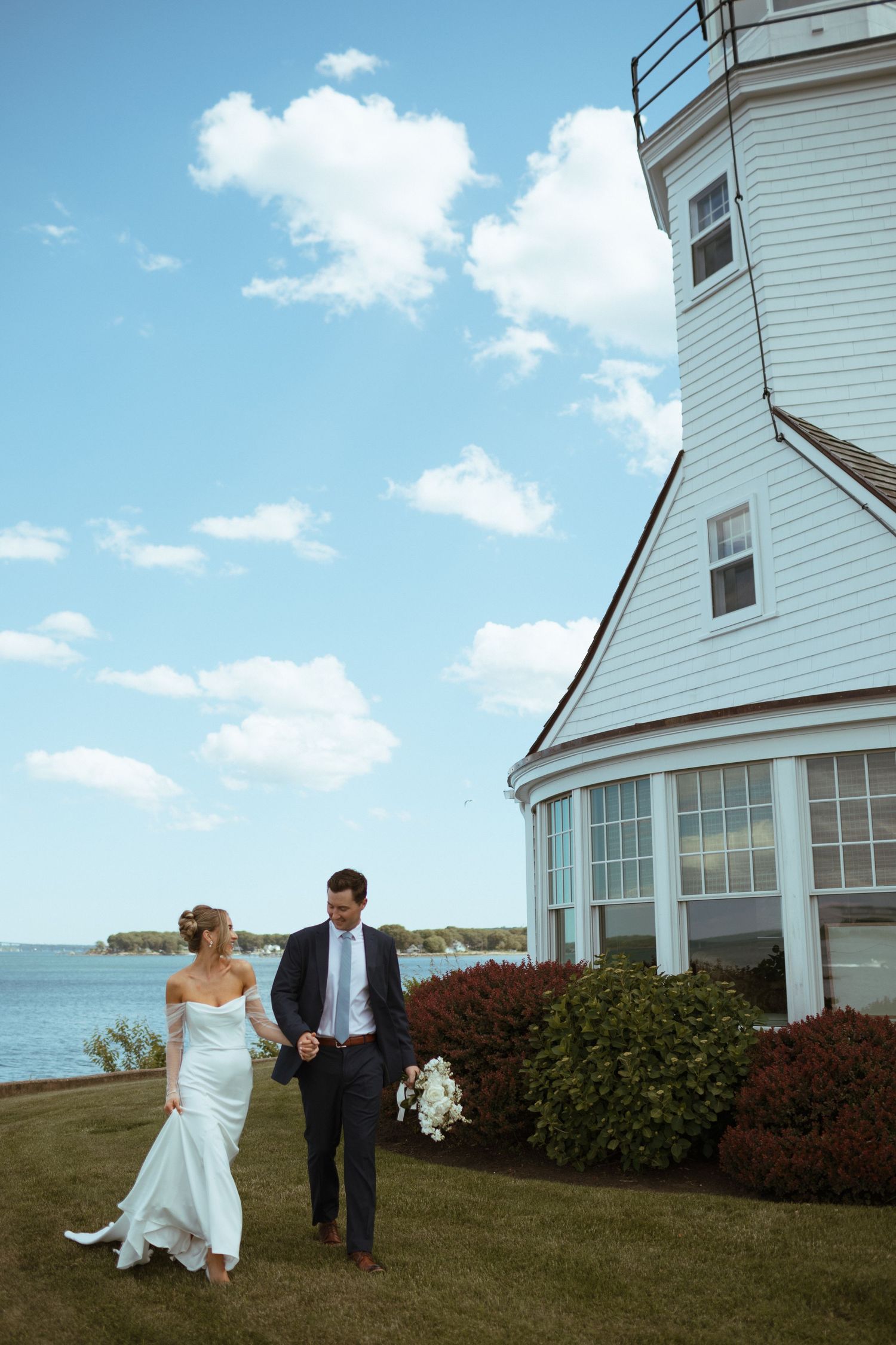 Bride and groom walk together beside a white lighthouse with ocean views under a bright blue sky.