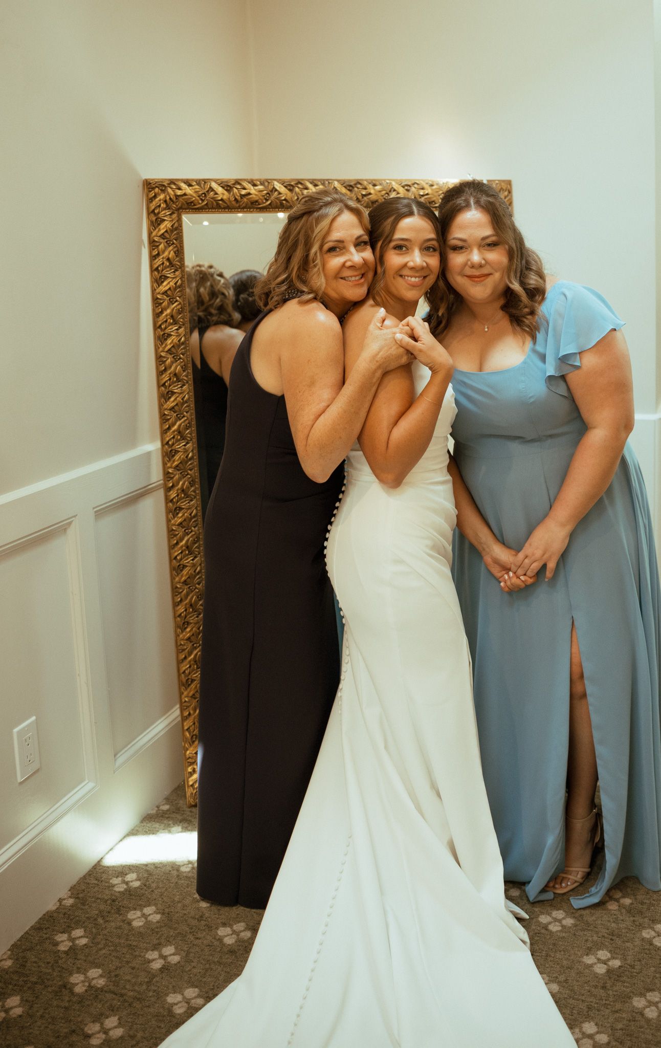 Bride in fitted white gown posing with two women near gold mirror before wedding.