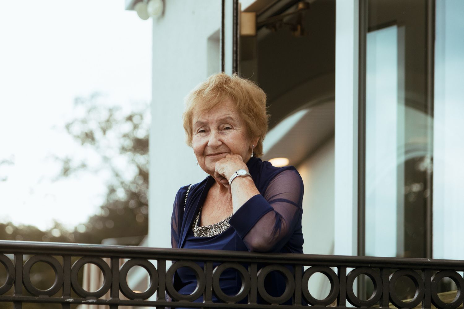 Elegant elderly woman smiling on a balcony, wearing a navy blue outfit.