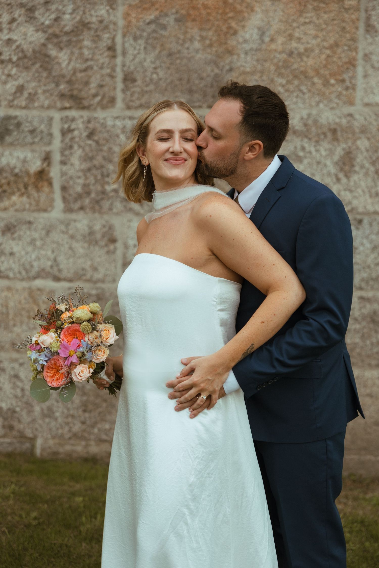 Groom in navy suit kisses bride on the cheek as she smiles holding a vibrant mixed floral bouquet against a stone wall.