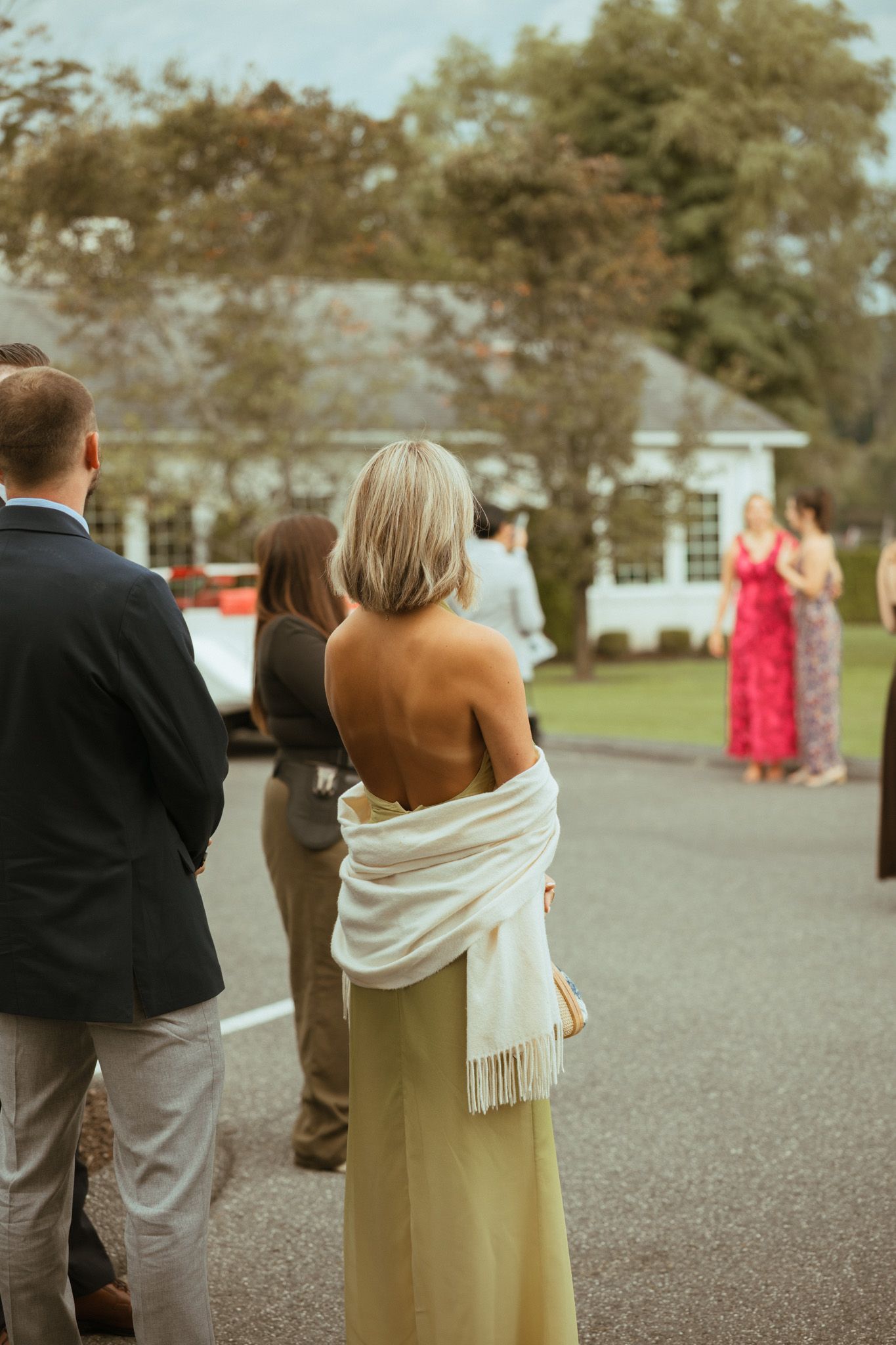 Wedding guest in a backless halter top with a white shawl standing among guests at an outdoor reception.