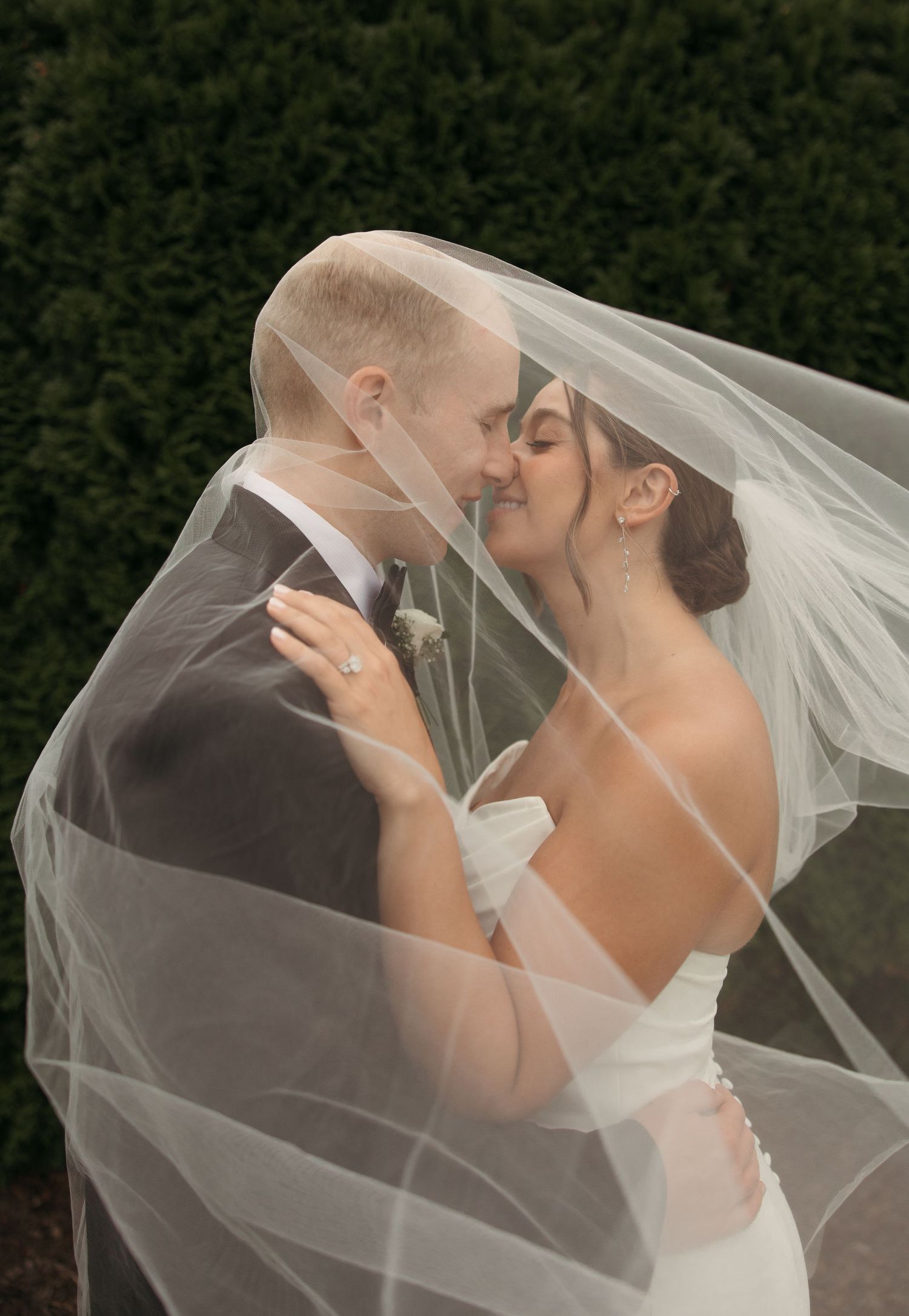 Bride and groom share a romantic kiss under flowing white veil against a lush green hedge backdrop.