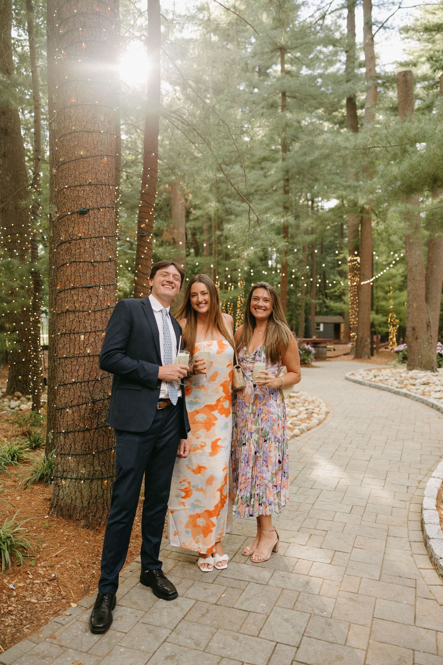 Three wedding guests posing on stone path surrounded by pine trees with string lights.