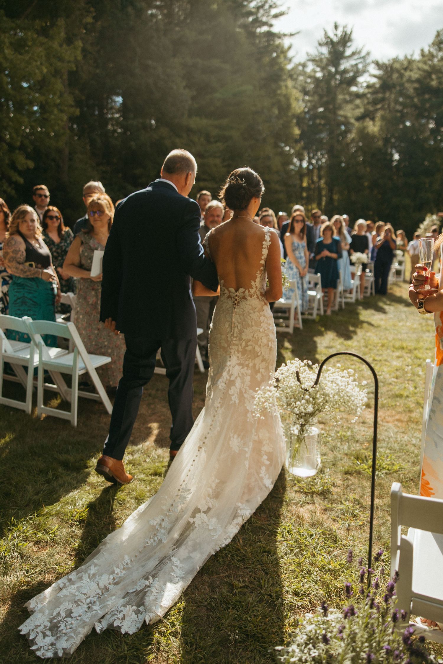 Bride in lace gown with long train walks down outdoor aisle with escort at sunny garden wedding ceremony.