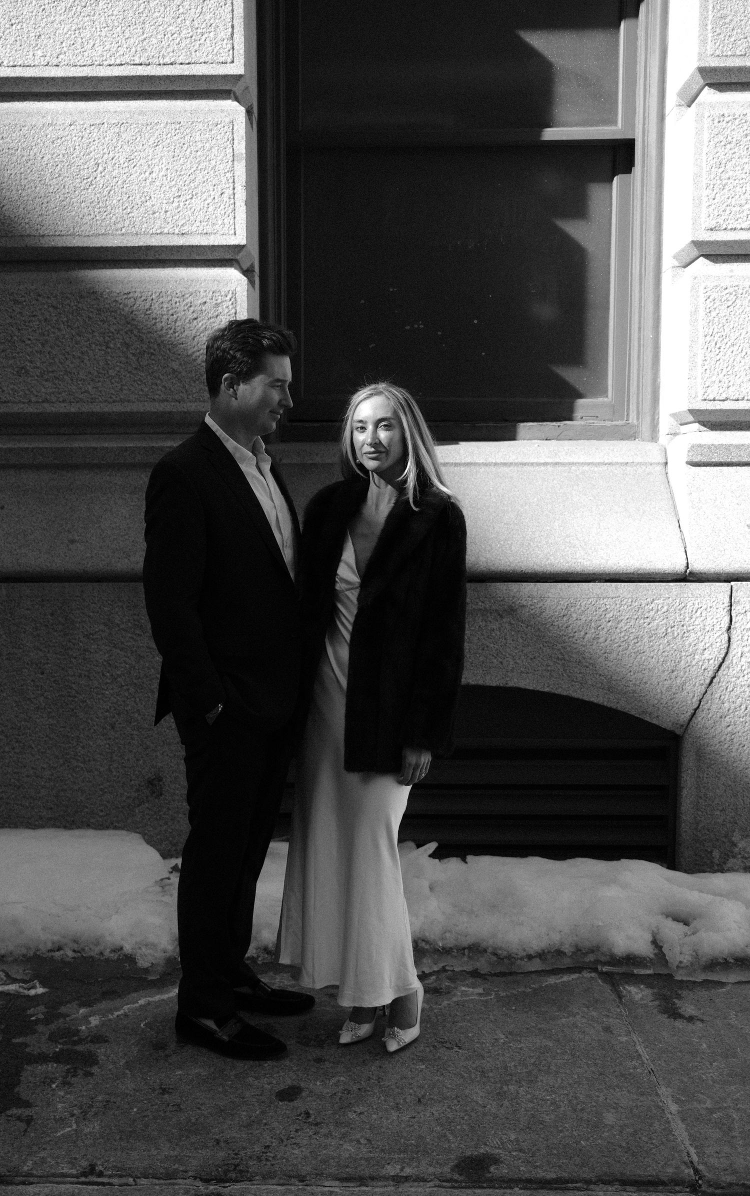 Black and white photo of an elegant couple standing together outside a stone building with snow on ground.film-inspired