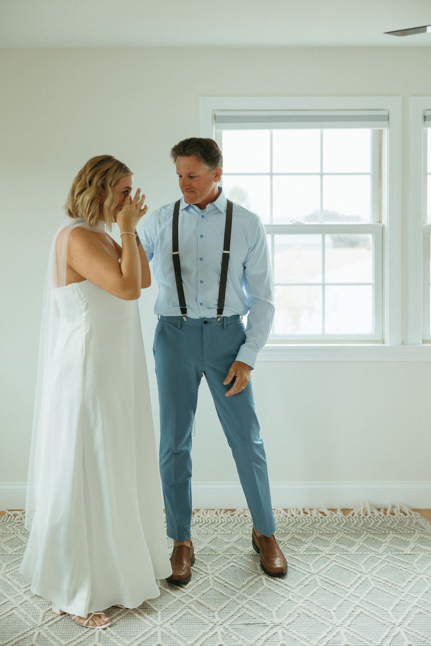 Emotional bride wipes tears during first look with groom in blue pants and suspenders by window.
