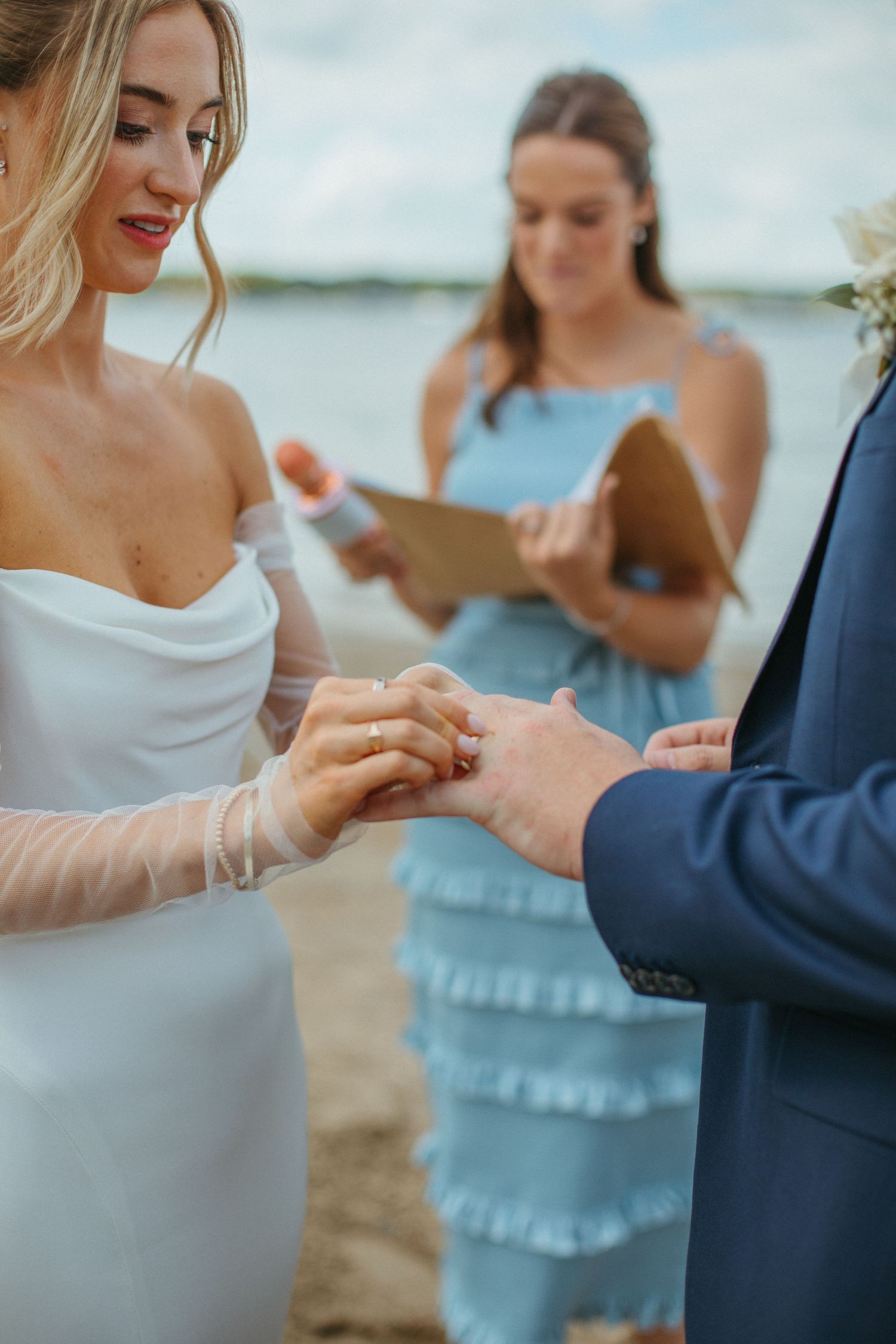 Bride placing gold wedding ring on groom's finger during beach ceremony with female officiant in blue dress.