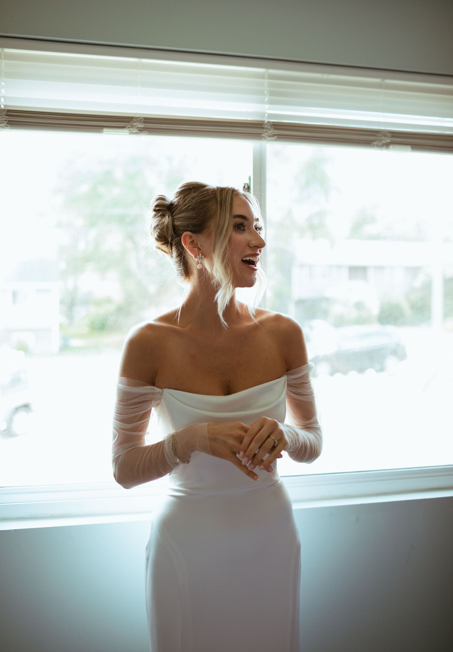 Radiant bride in off-shoulder long sleeve white wedding gown laughing by bright window before ceremony.