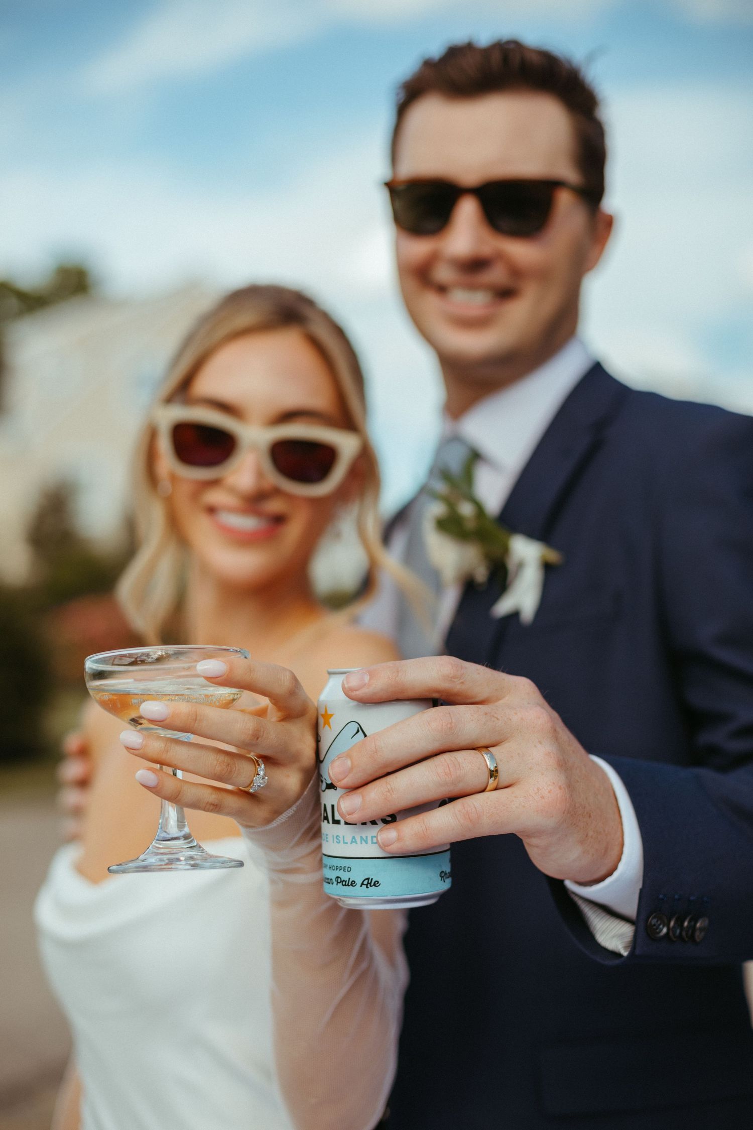 Smiling bride and groom in sunglasses toasting with a cocktail glass and beer can on their wedding day.