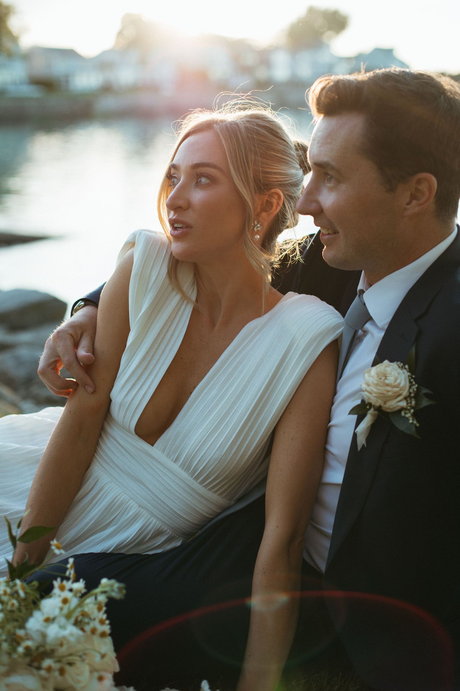 Bride in deep V-neck white gown and groom in dark suit sitting by waterfront at golden hour.
