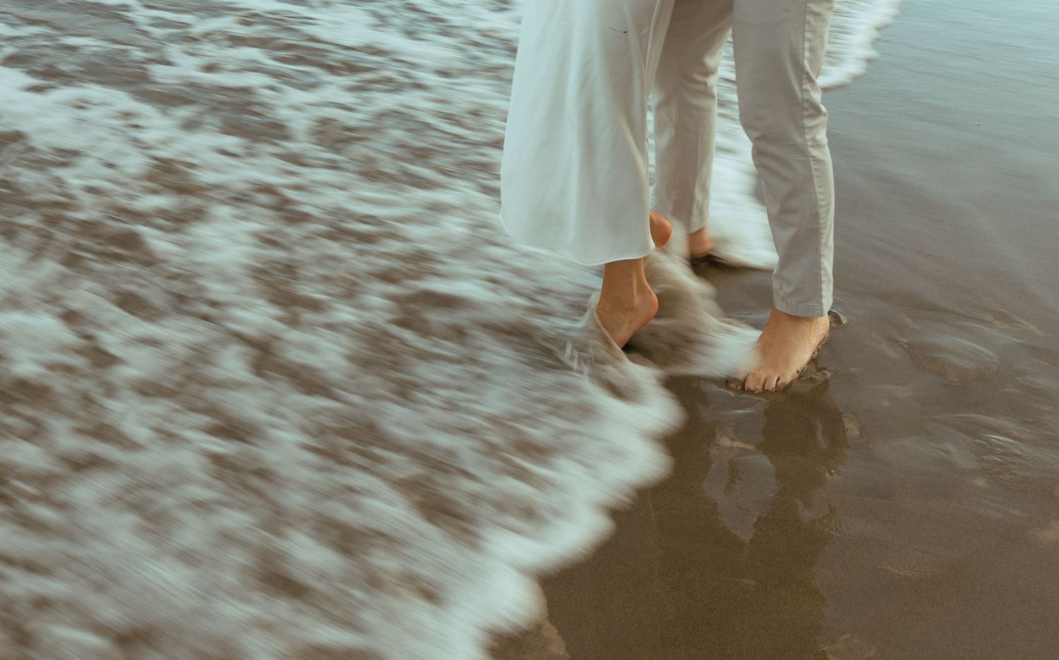 Romantic couple standing barefoot in ocean waves on a beach, captured in a dreamy wedding photography style.