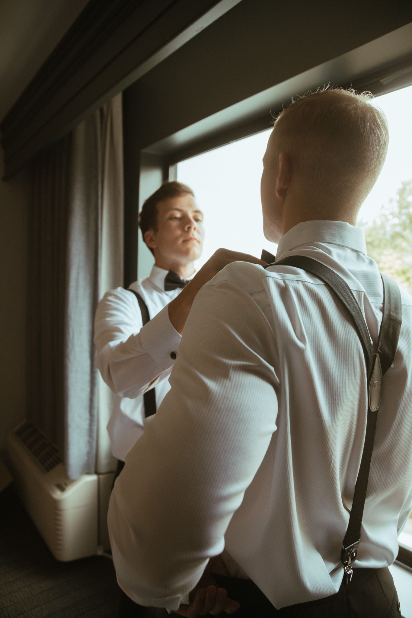 Groomsman helping groom adjust bow tie while getting ready near window on wedding day.