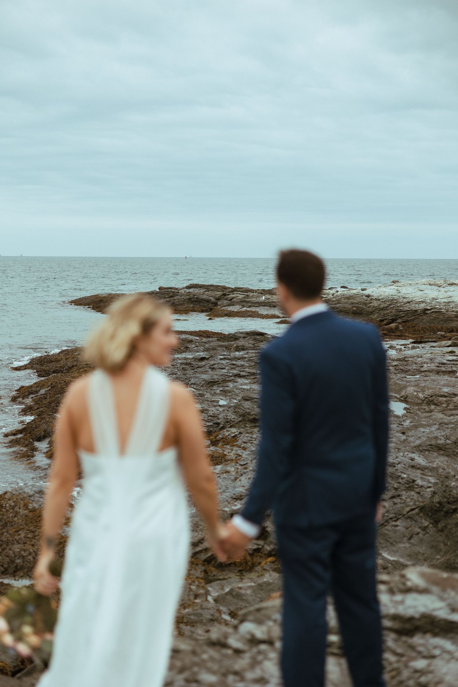Defocused artistic shot of bride and groom holding hands facing the rocky ocean shoreline on their wedding day.