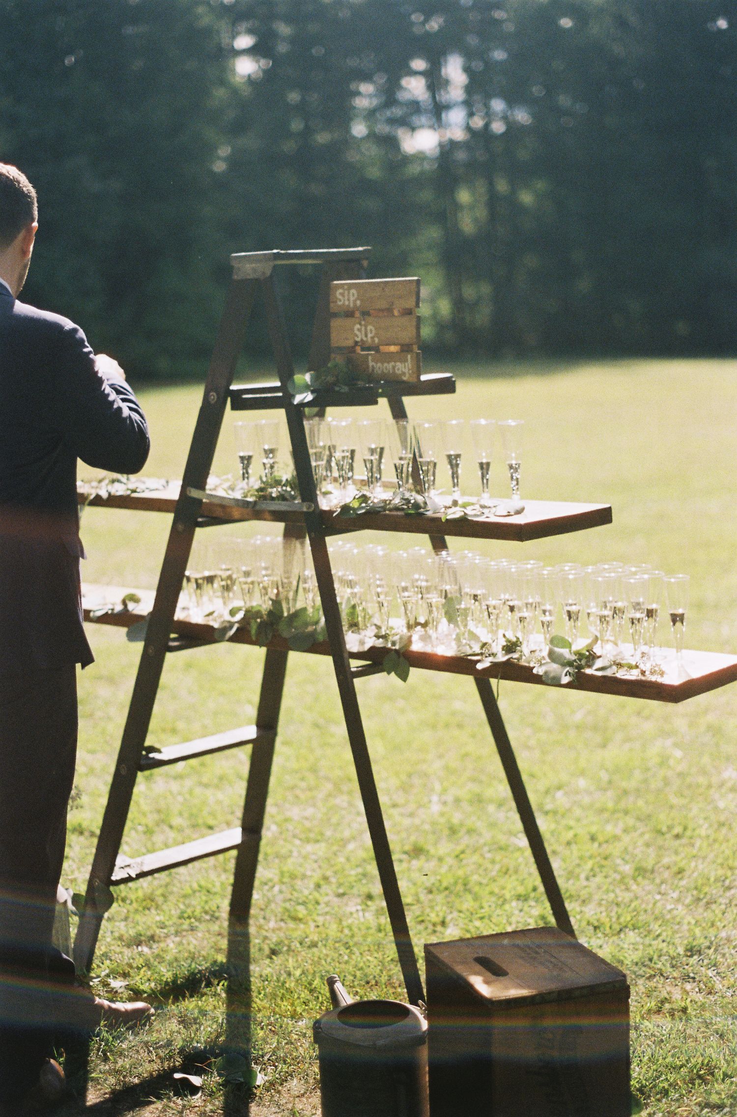 Champagne glasses displayed on rustic wooden ladder at outdoor wedding reception.film-inspired