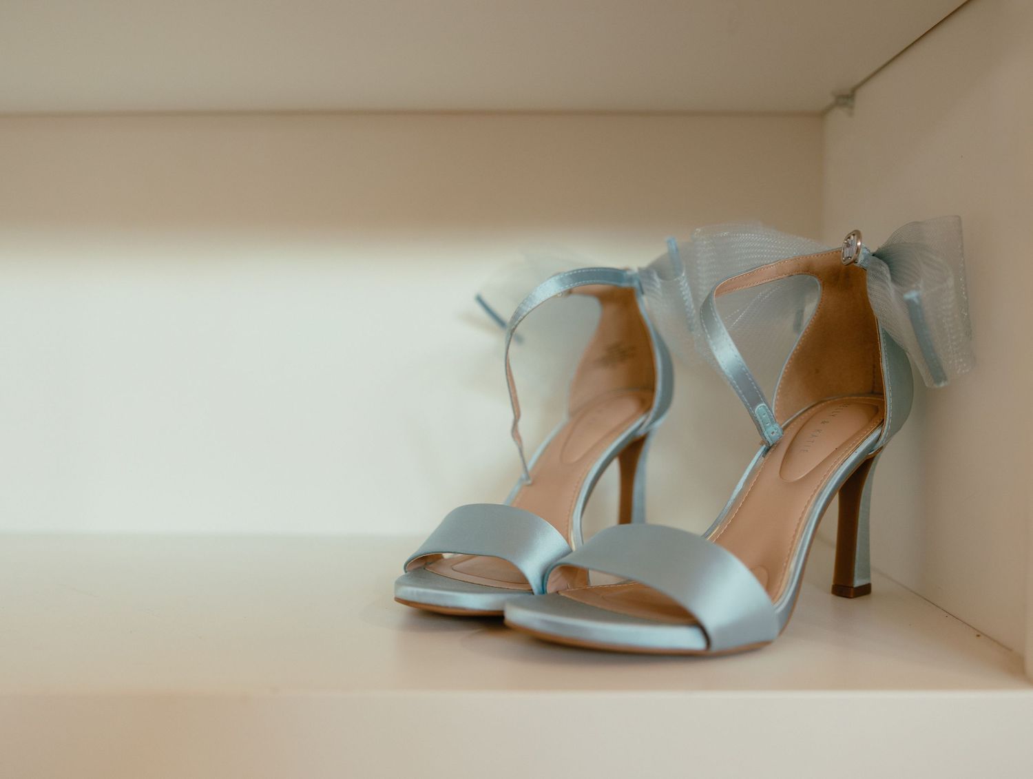 Elegant light blue satin bridal heels with tulle bow detail displayed on white shelf.