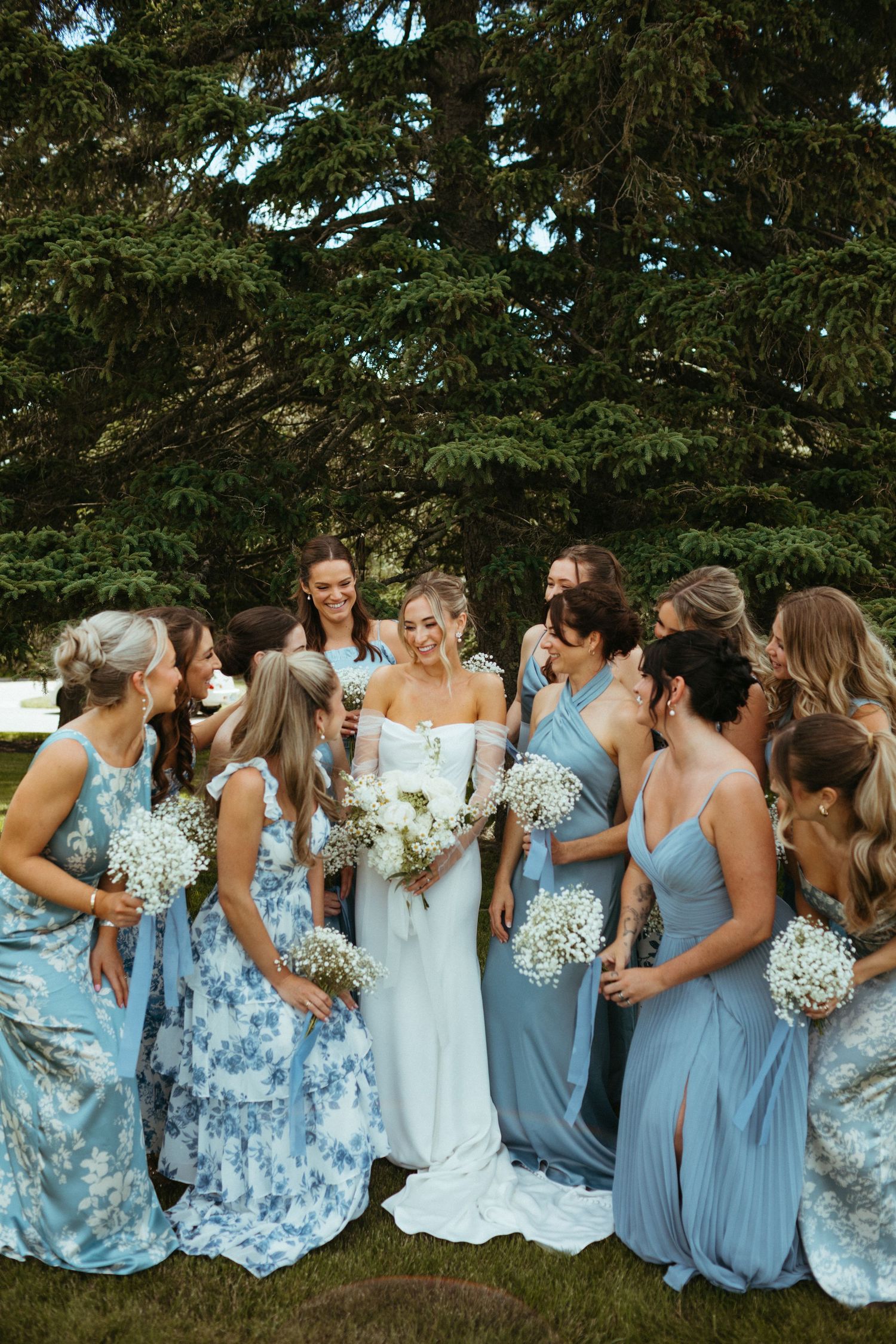 Bride in white off-shoulder gown surrounded by bridesmaids in blue dresses holding baby's breath bouquets.