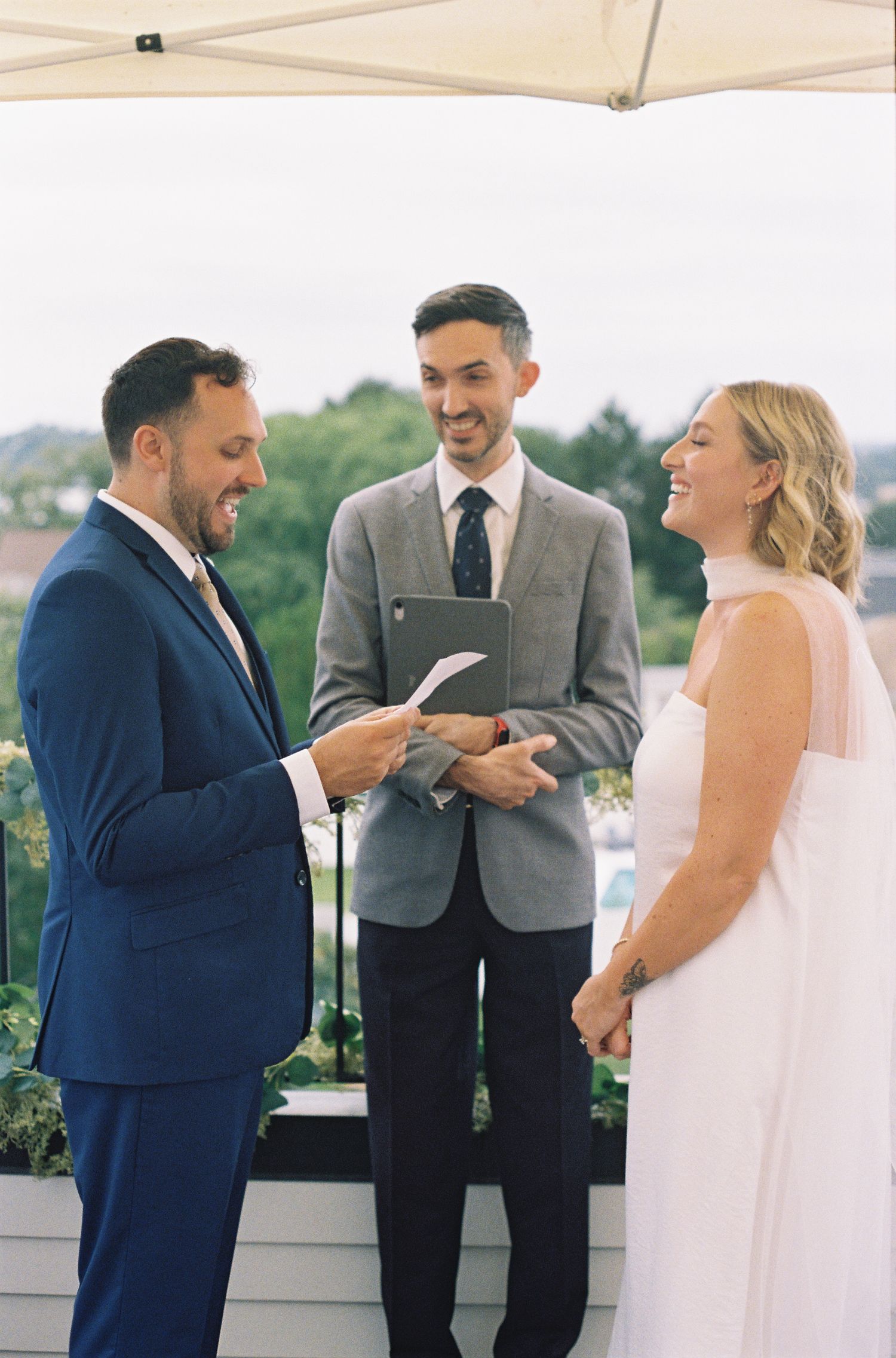 Bride and groom laugh joyfully during outdoor rooftop wedding ceremony while officiant reads from tablet.