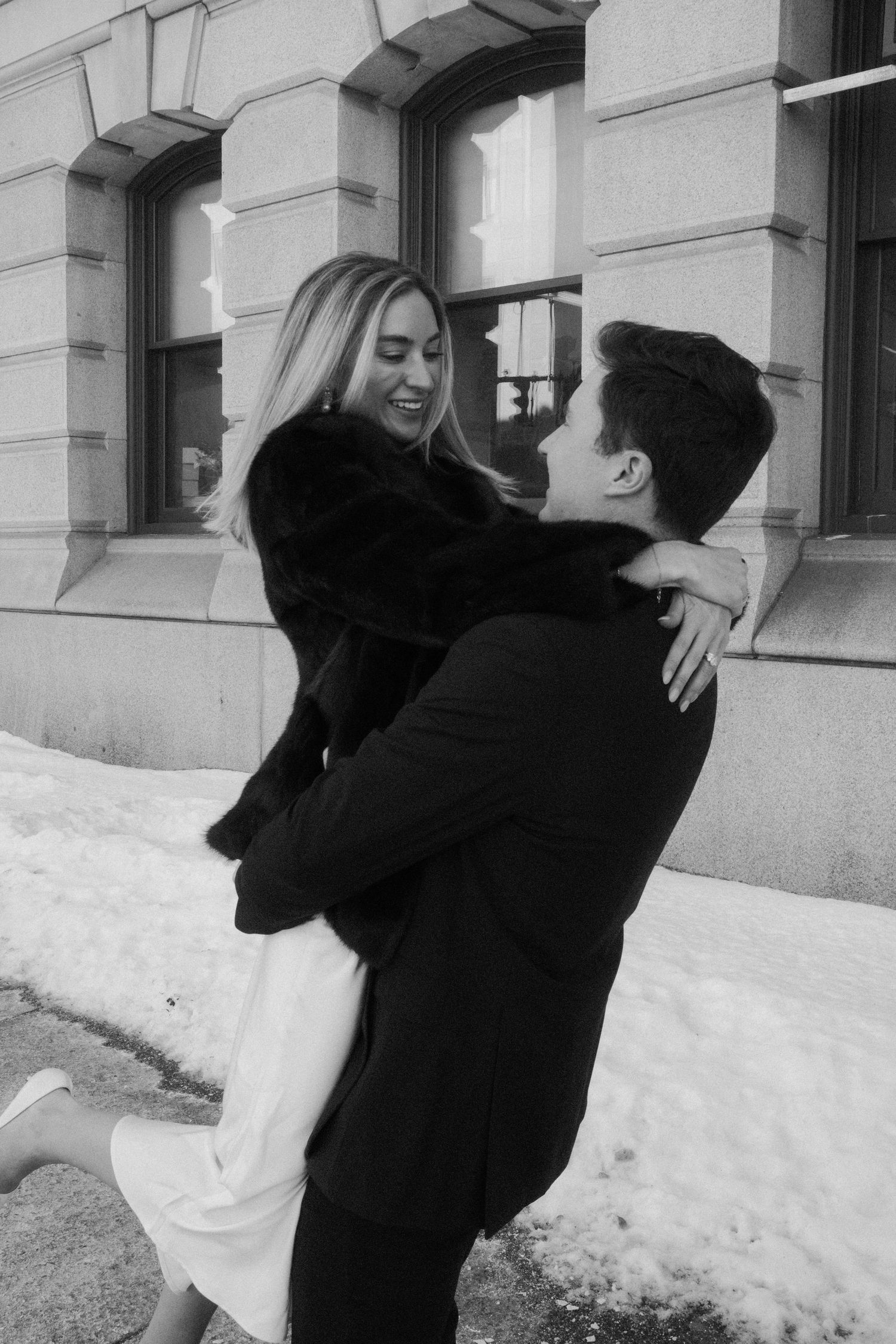 Black and white photo of a laughing woman being lifted by her partner outside a snowy urban building.film-inspired