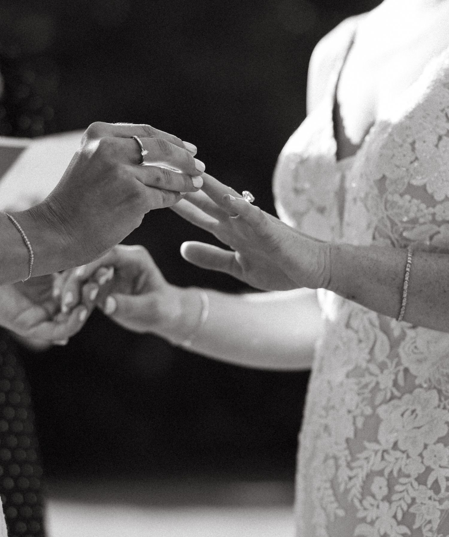 Black and white close-up of two brides exchanging wedding rings during outdoor ceremony.