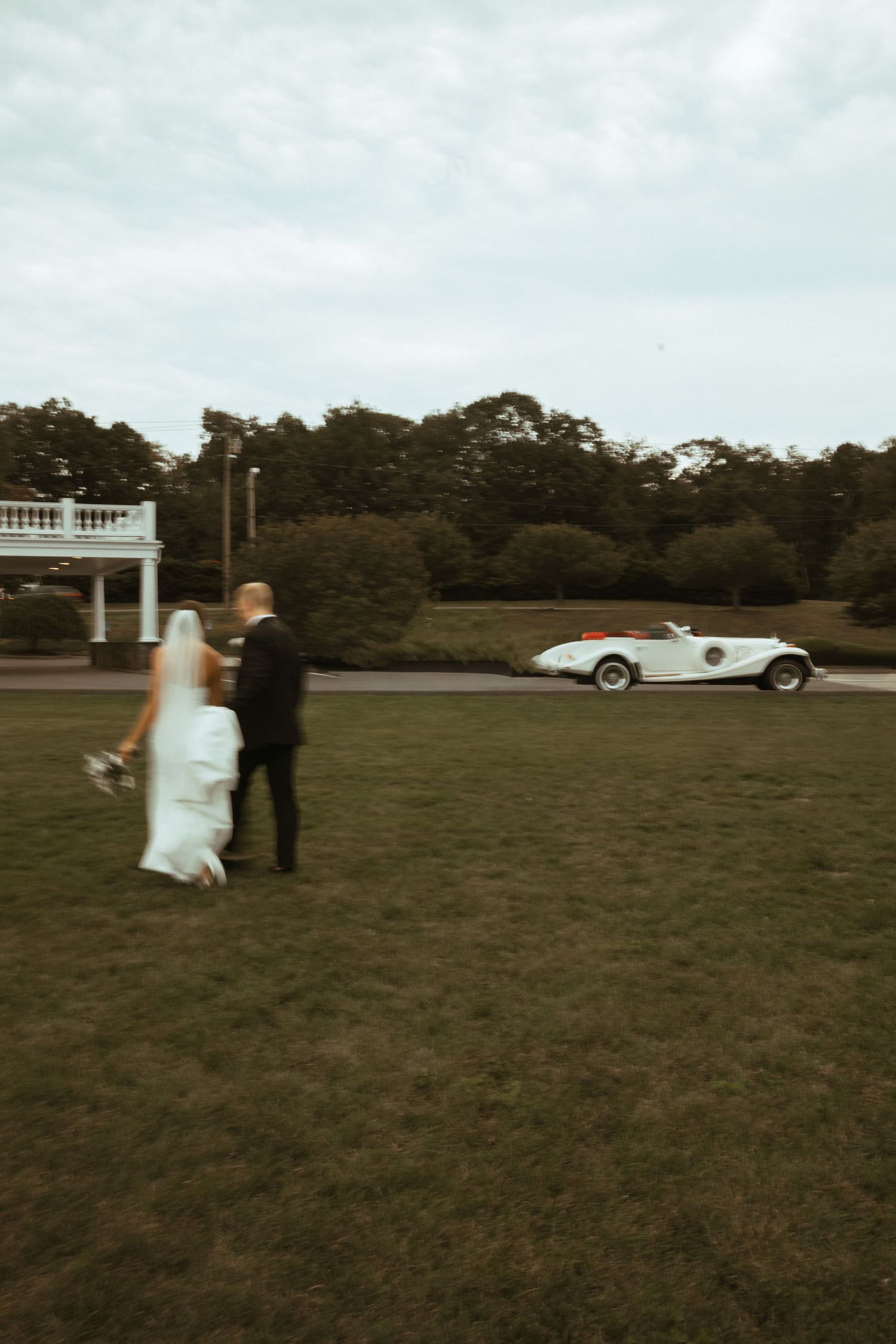 Bride and groom walking toward a vintage white convertible car on a green lawn at their wedding venue.