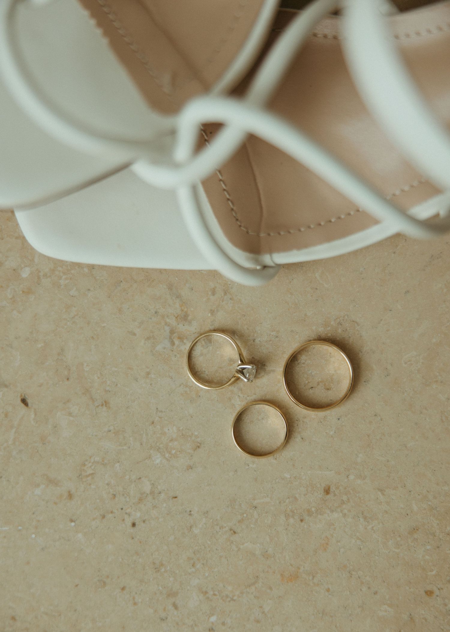 Gold wedding rings and engagement ring displayed beside white bridal heels on stone surface.