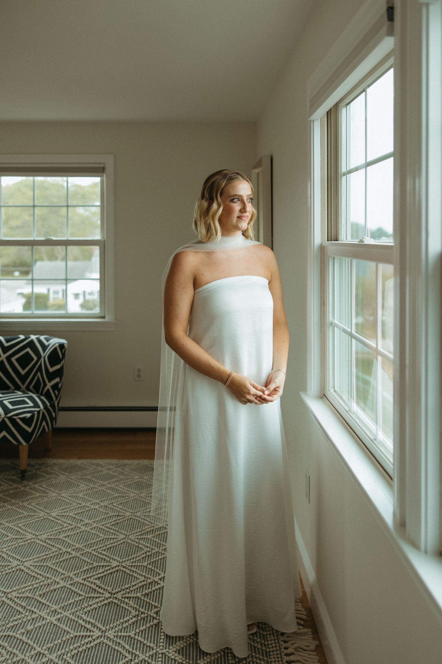 Beautiful bride in strapless white wedding gown with veil gazes out window in natural light.