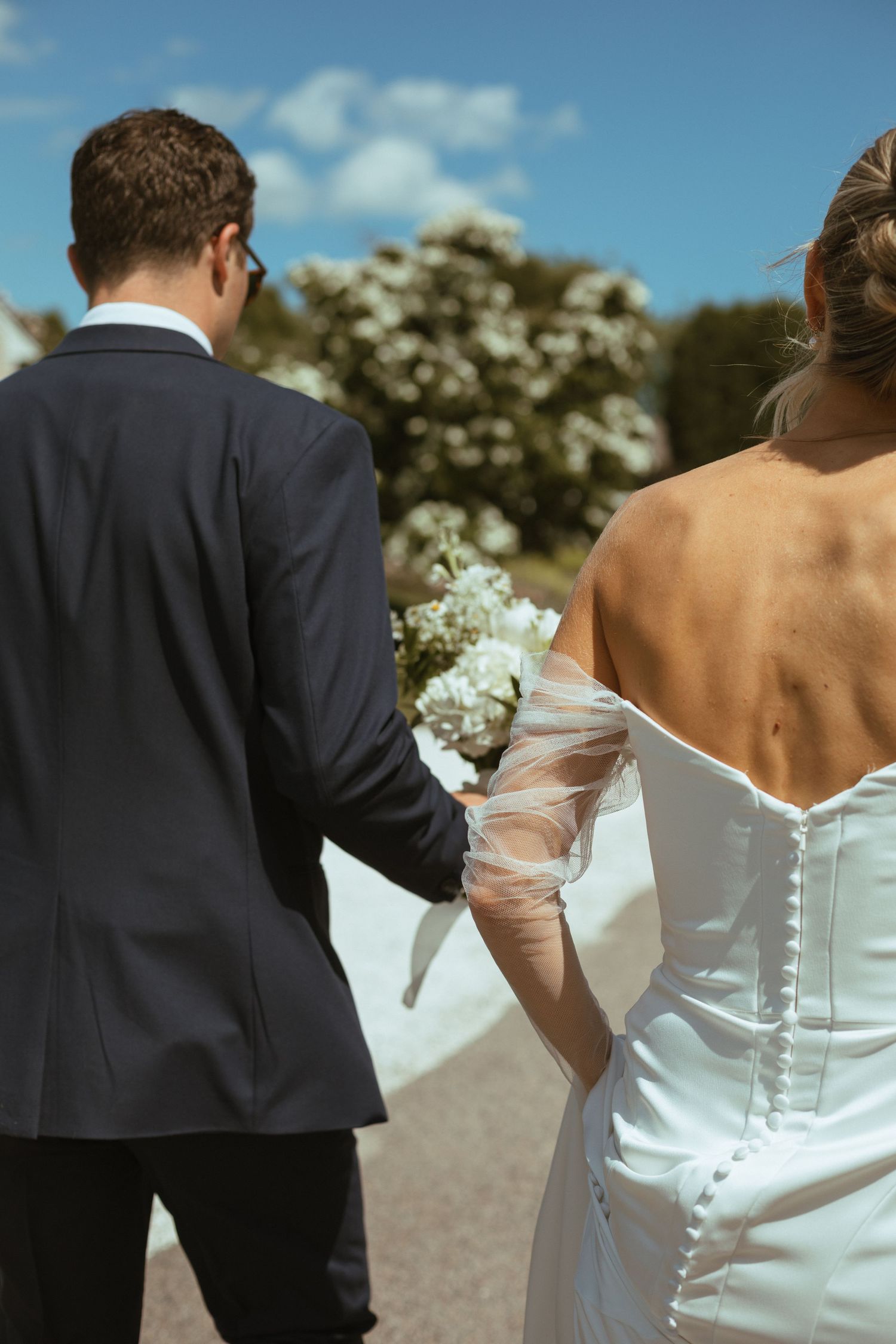 Bride and groom walk hand in hand, bride in backless button-up gown with tulle gloves holding white bouquet.