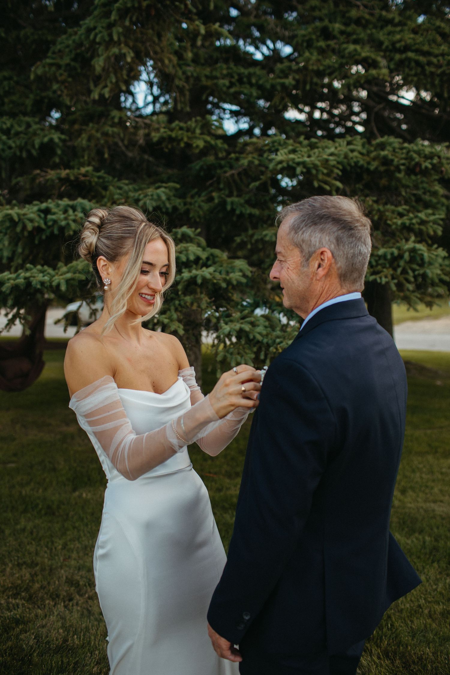 Bride in white off-shoulder gown pinning boutonniere on older man in navy suit near evergreen trees.