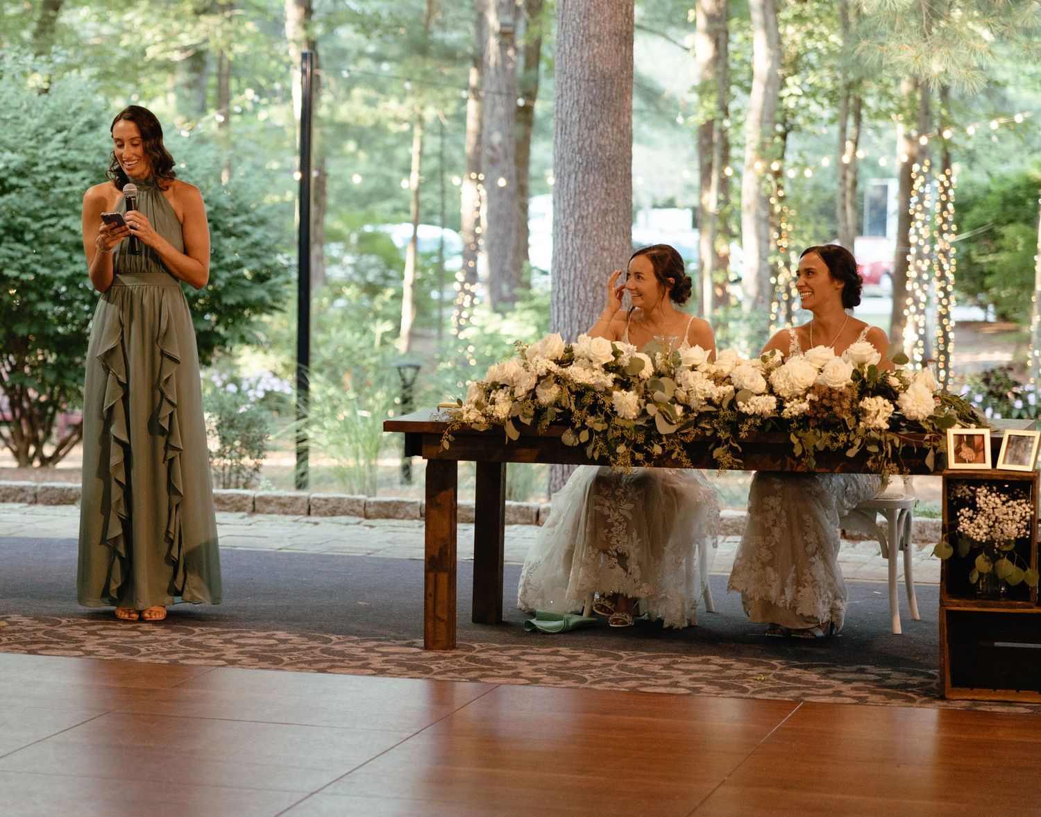 Bridesmaid in sage dress giving speech while two brides laugh at floral sweetheart table.film-inspired
