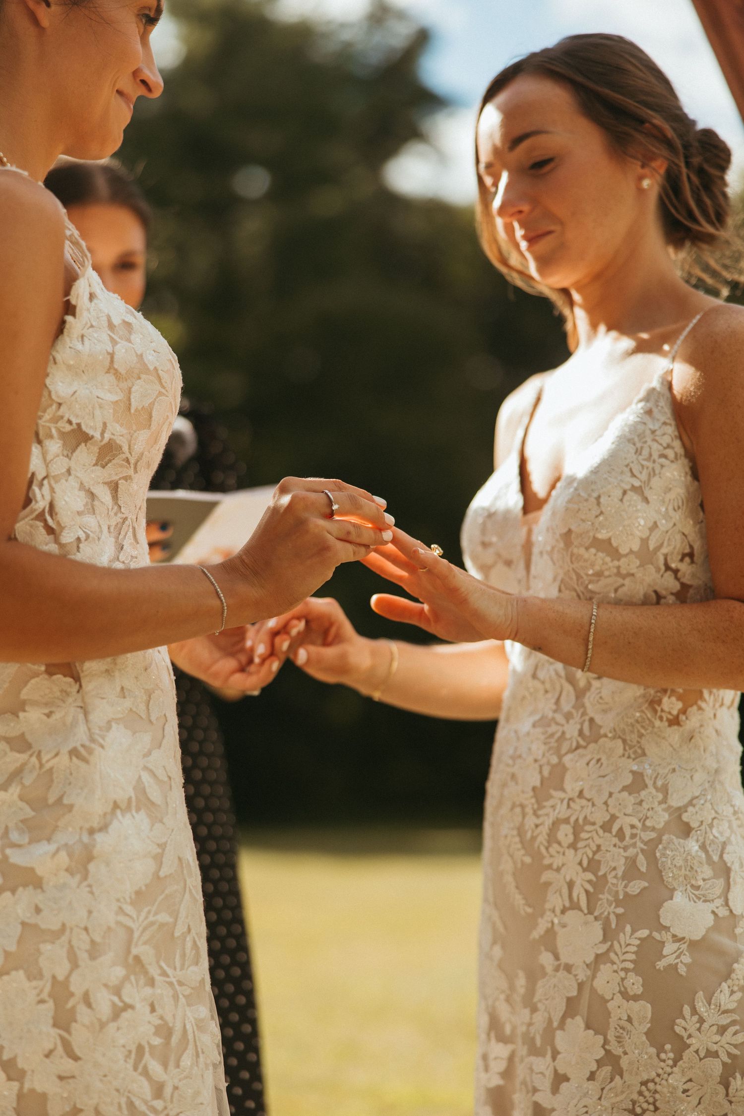Two brides exchange wedding rings in warm golden sunlight during intimate outdoor ceremony.