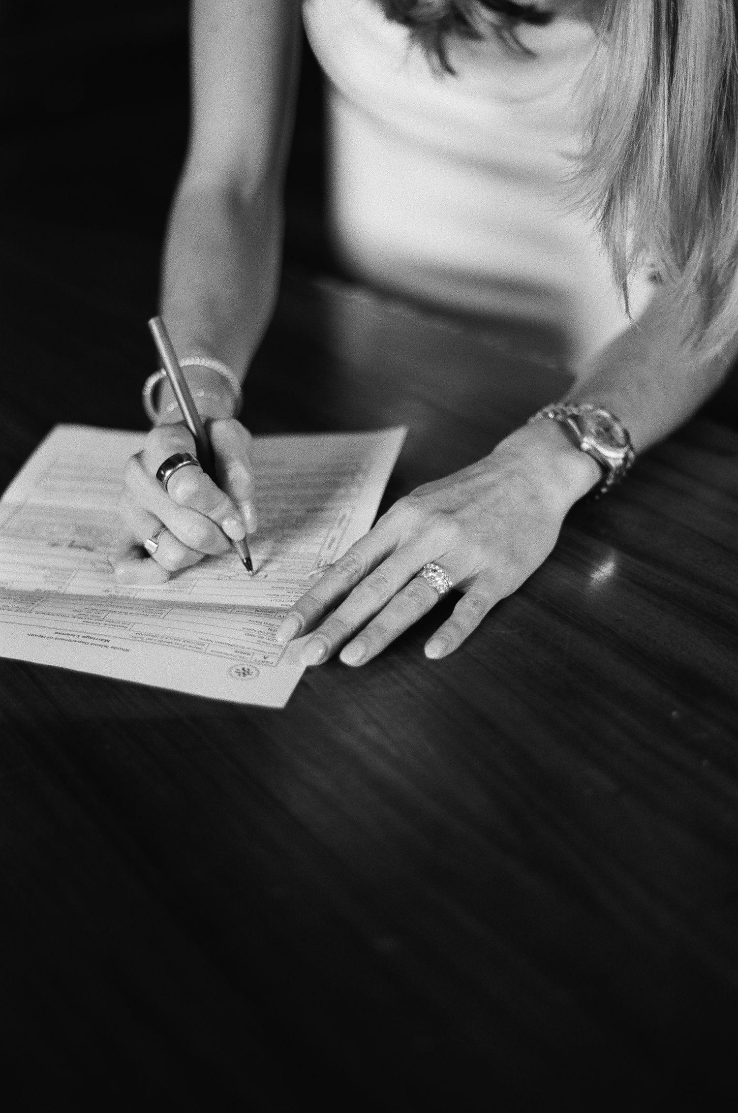 Close-up of bride signing marriage certificate with pen, wearing bracelet and rings at wooden table.film-inspired