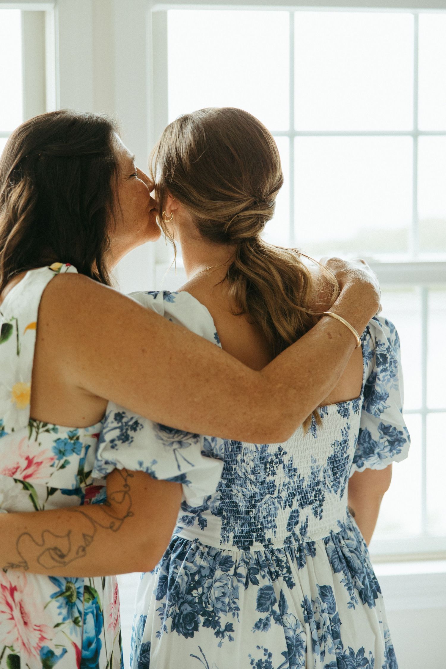 Two women in floral dresses sharing an emotional embrace before wedding ceremony.