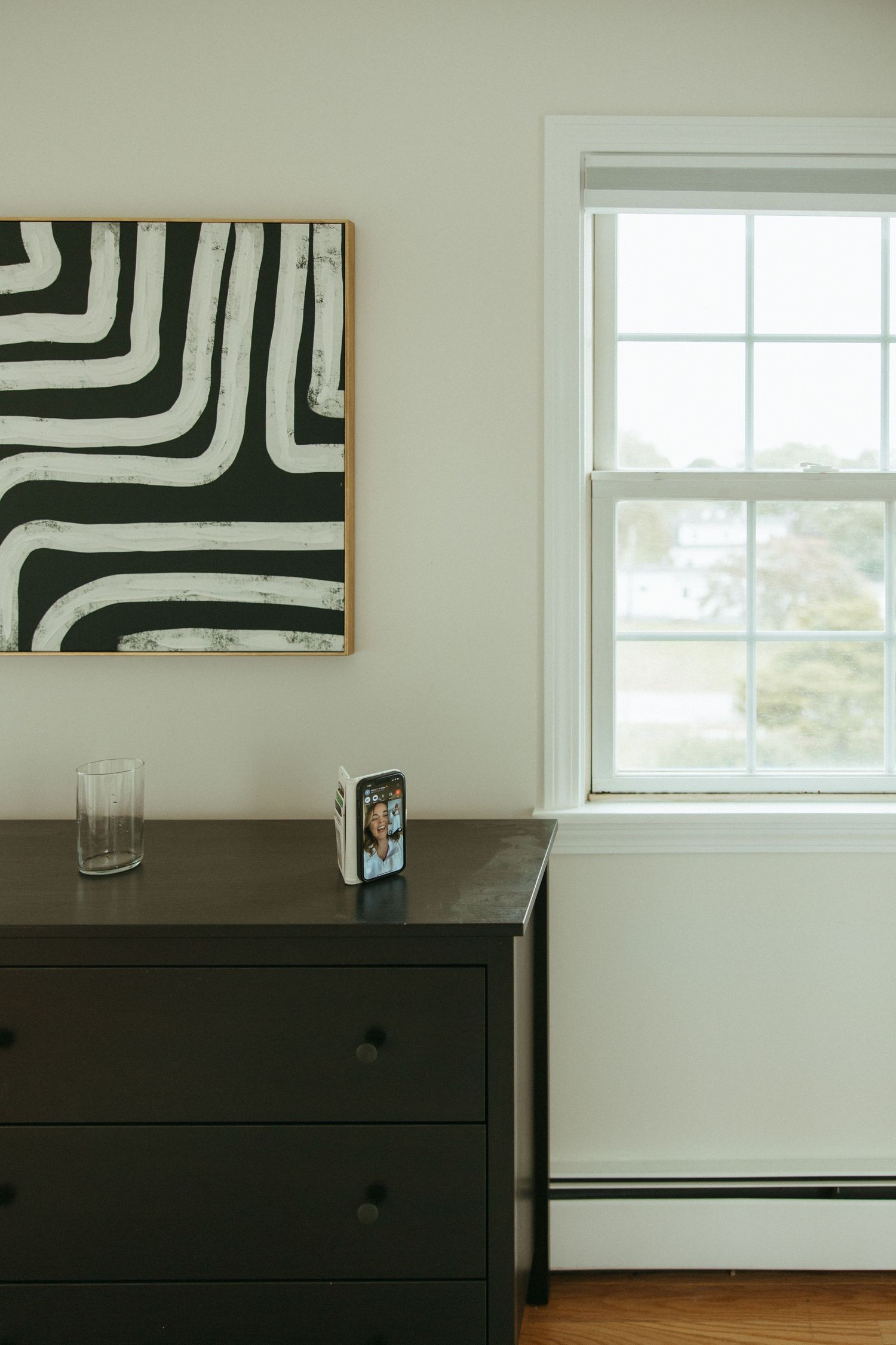 Black dresser with framed photo and glass beside bold black and white abstract art near window.
