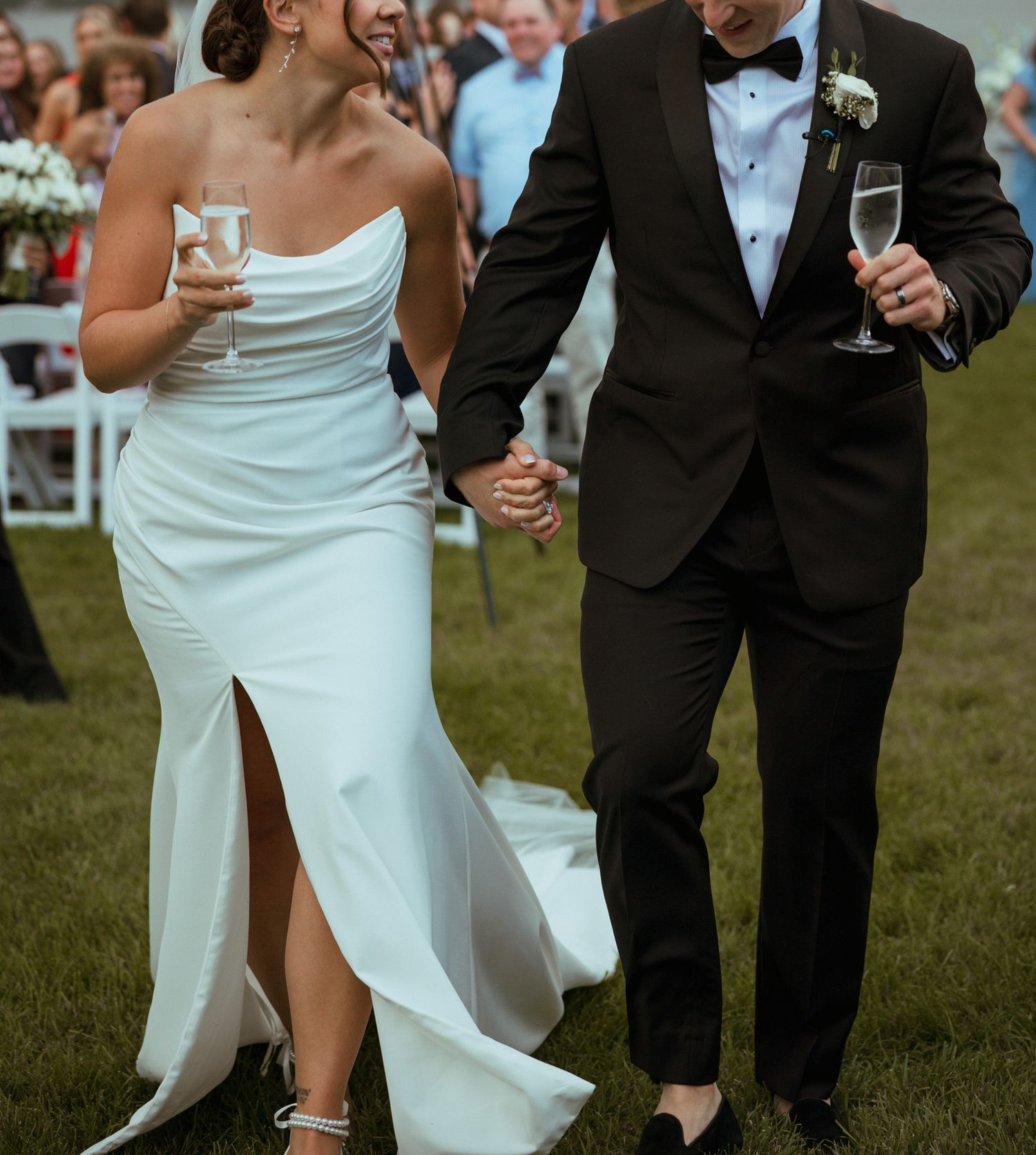 Newlyweds walk hand in hand holding champagne glasses celebrating after outdoor wedding ceremony.