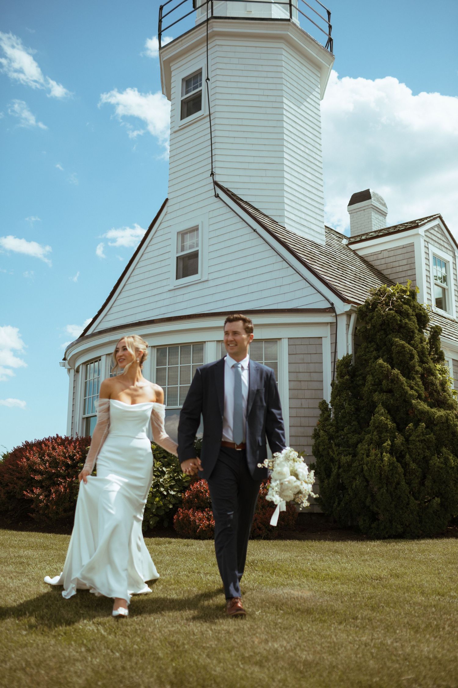 Bride and groom walking hand in hand on lawn beside white lighthouse on sunny day.