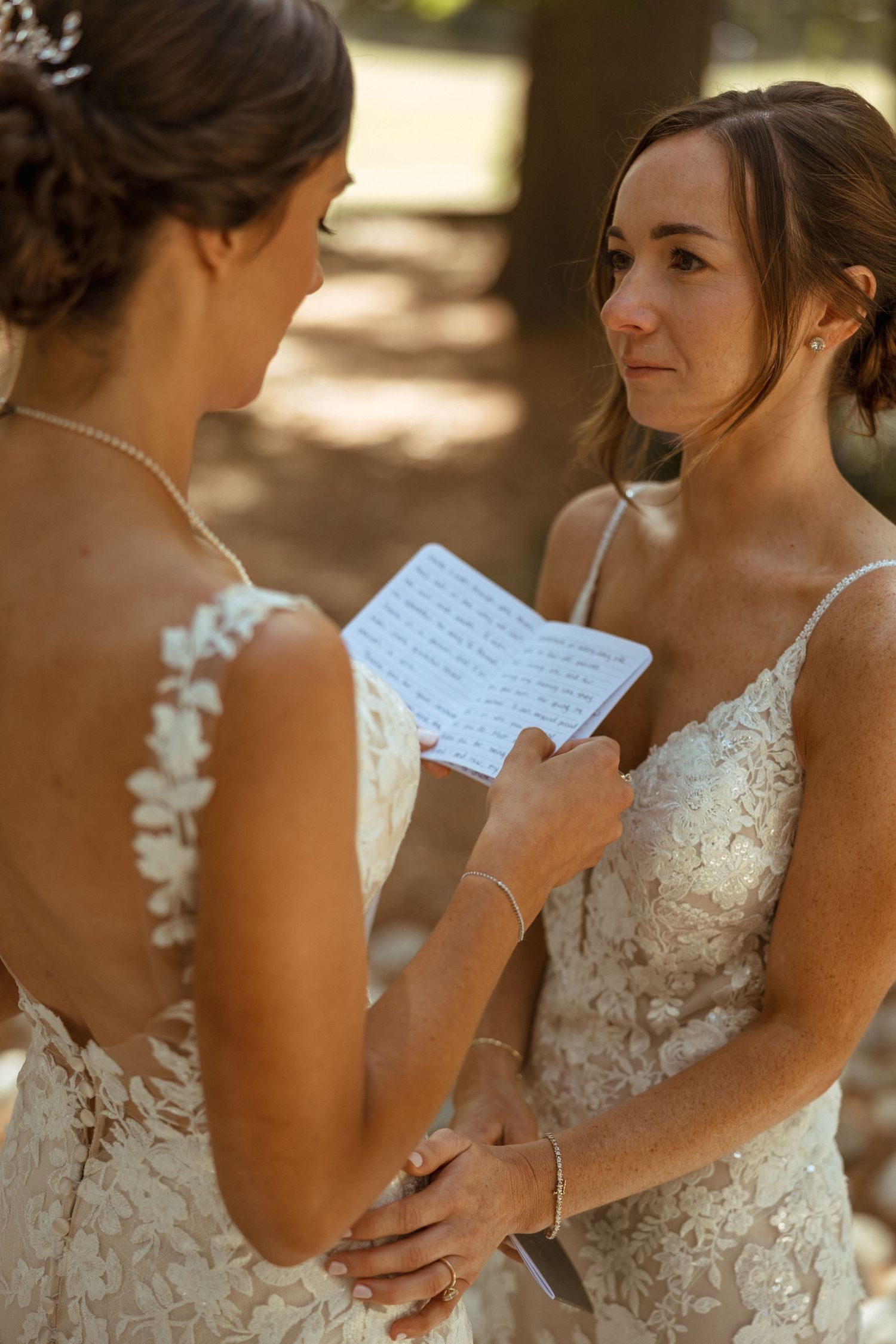 Two brides in lace gowns exchange emotional handwritten vows while holding hands at outdoor wedding.