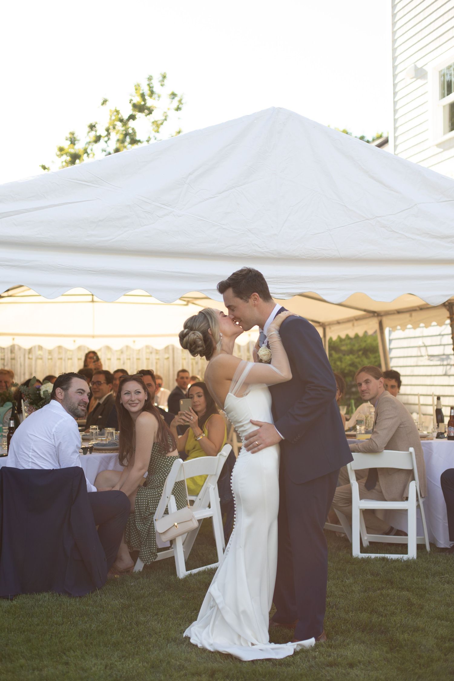 Newlyweds kissing under white reception tent while guests celebrate at outdoor garden wedding.