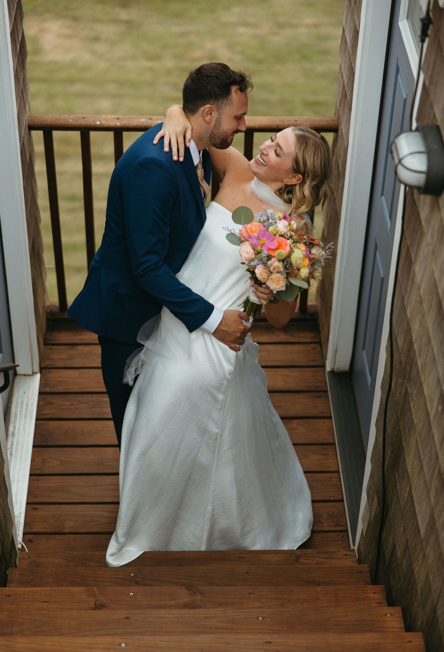Bride and groom share a romantic moment on wooden stairs, she holds a colorful bouquet in a white gown, he wears a navy suit.