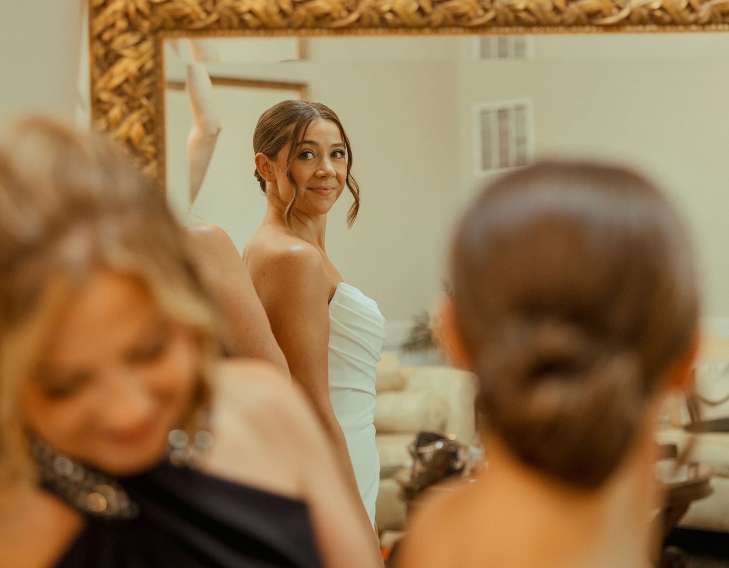 Bride smiling in mirror reflection while bridesmaids help her prepare for wedding day.