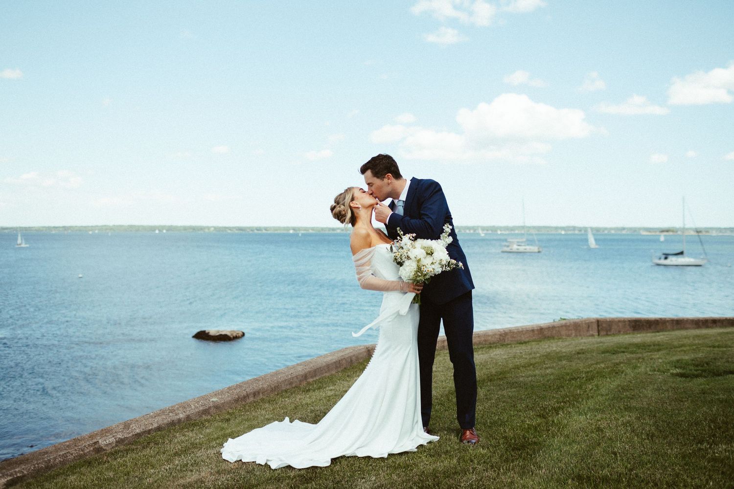 Bride and groom share a romantic kiss on a grassy cliff overlooking a sparkling blue harbor with sailboats.