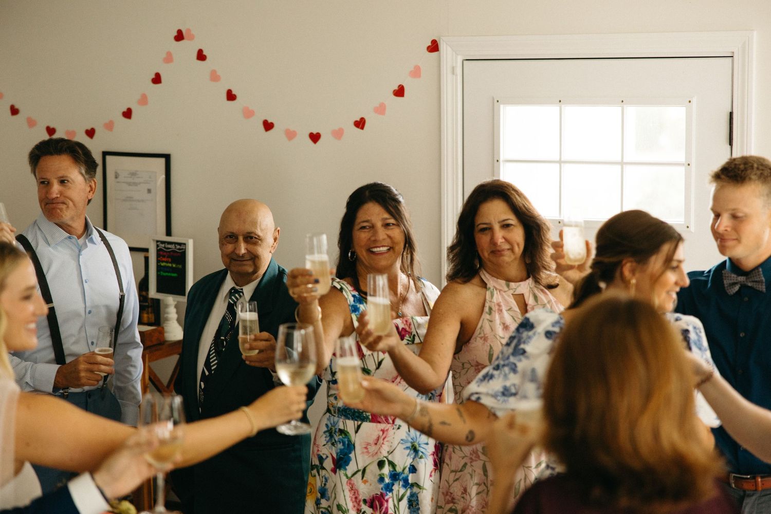 Wedding guests raise champagne glasses in a celebratory toast indoors beneath red heart garland.