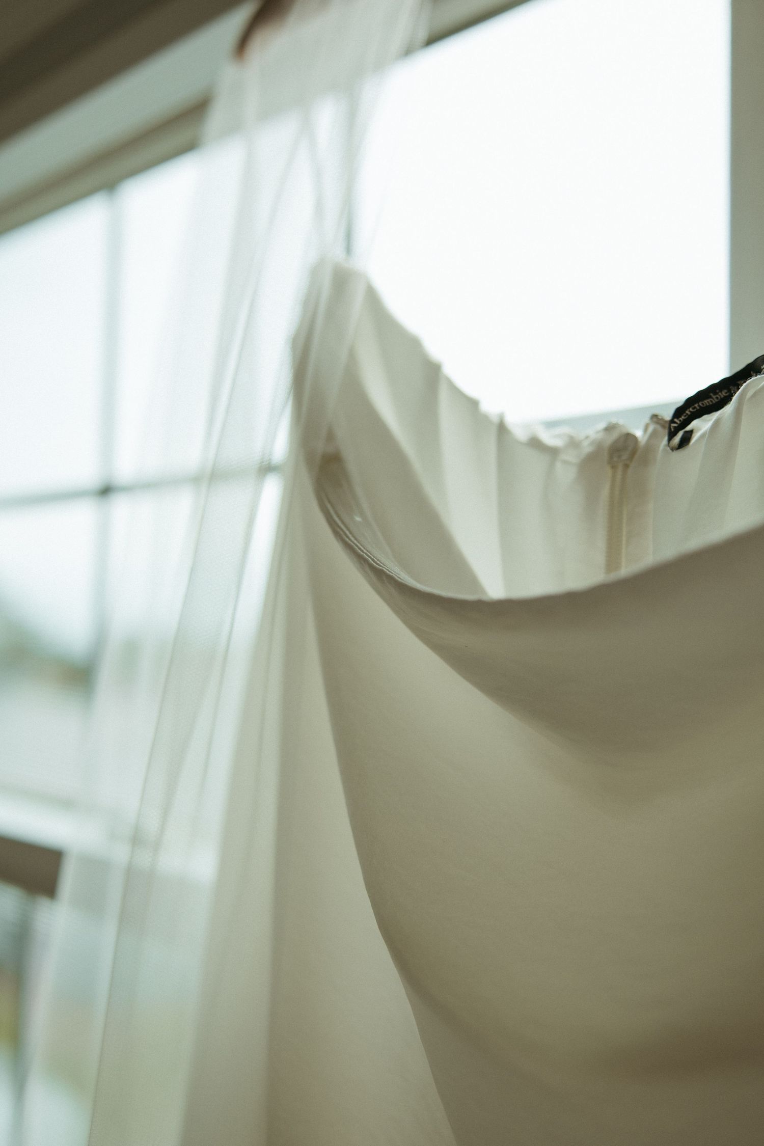 White wedding dress with veil hanging near bright window before ceremony.