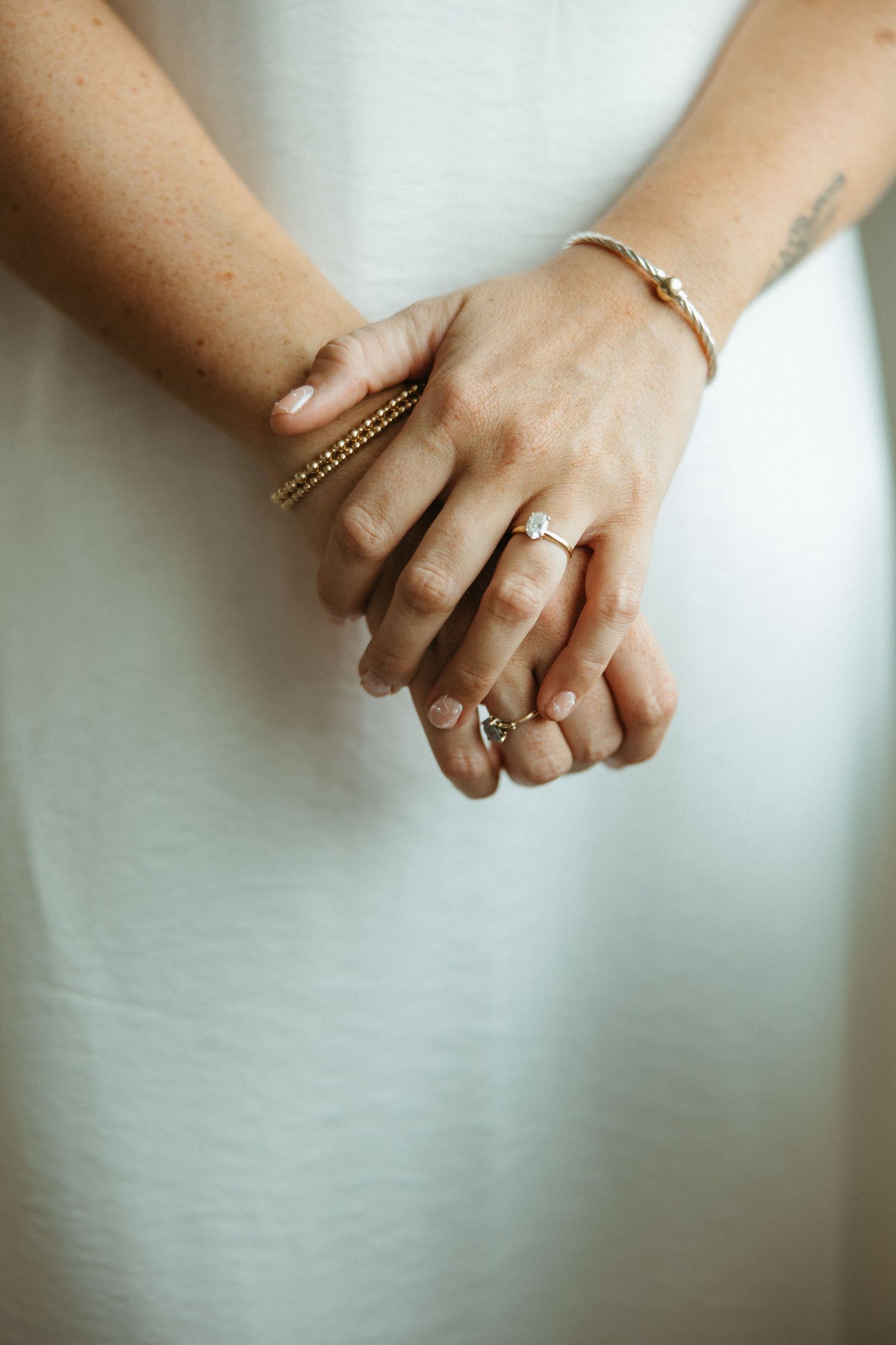 Close-up of bride's hands clasped together wearing engagement ring and gold bracelet on white gown.