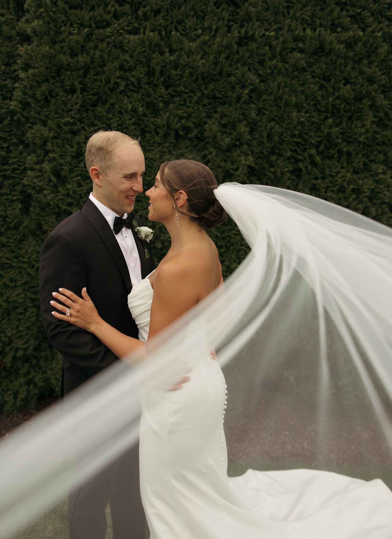 Bride and groom gaze lovingly at each other as wedding veil flows dramatically in the breeze outdoors.